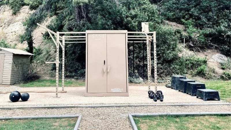 Outdoor fitness station with pull-up bars, kettlebells, medicine balls, and plyometric boxes in a grassy area surrounded by trees.