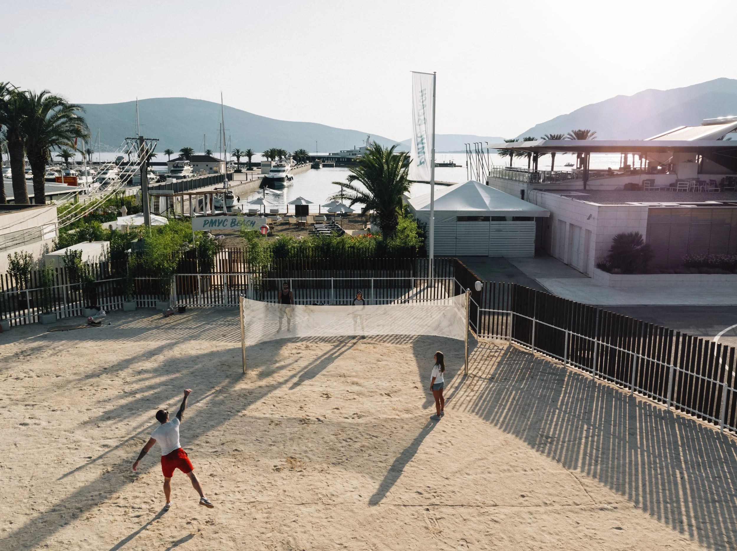 People playing beach volleyball at a sandy court near a marina with boats and yachts, surrounded by palm trees and hills in the background.