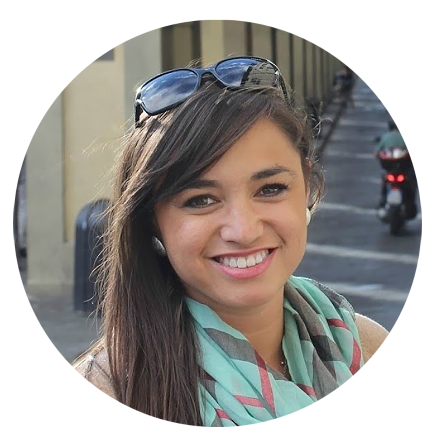 A picture of Amanda Sue, she has long brown hair, held back with sunglasses, smiles and wears a light blue and pink scarf around her neck. There's a street with a scooter parked in the back.