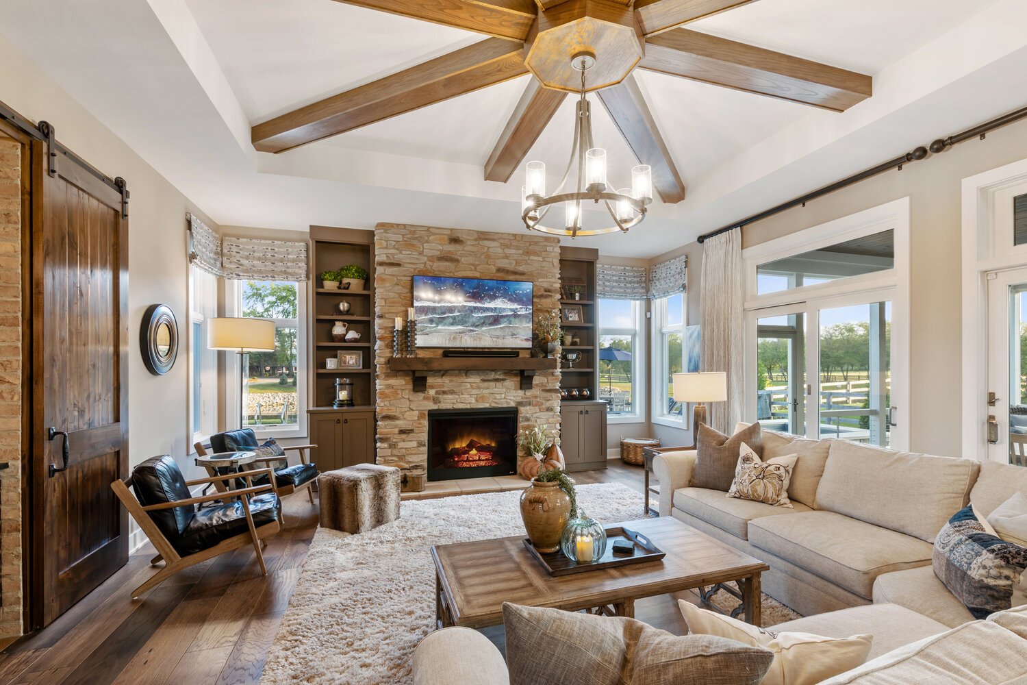 Open concept living area featuring wooden beams, stone fireplace, large windows, and comfortable furniture.