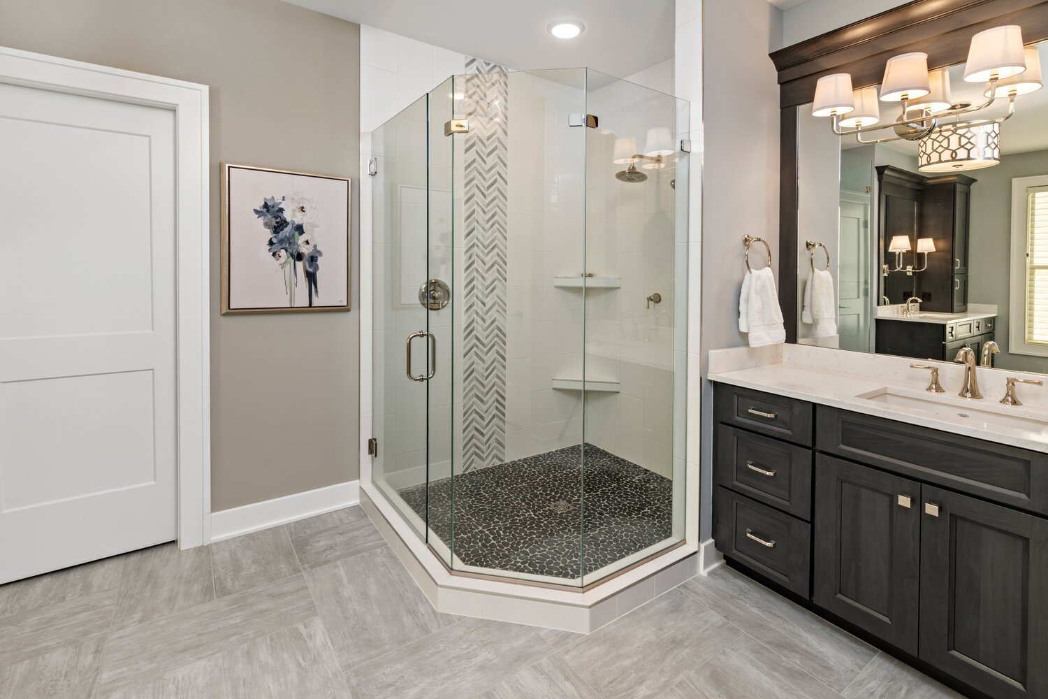 A luxurious bathroom showcasing a walk-in tiled shower with chevron accent wall, double vanity with dark wood cabinetry, and decorative lighting.