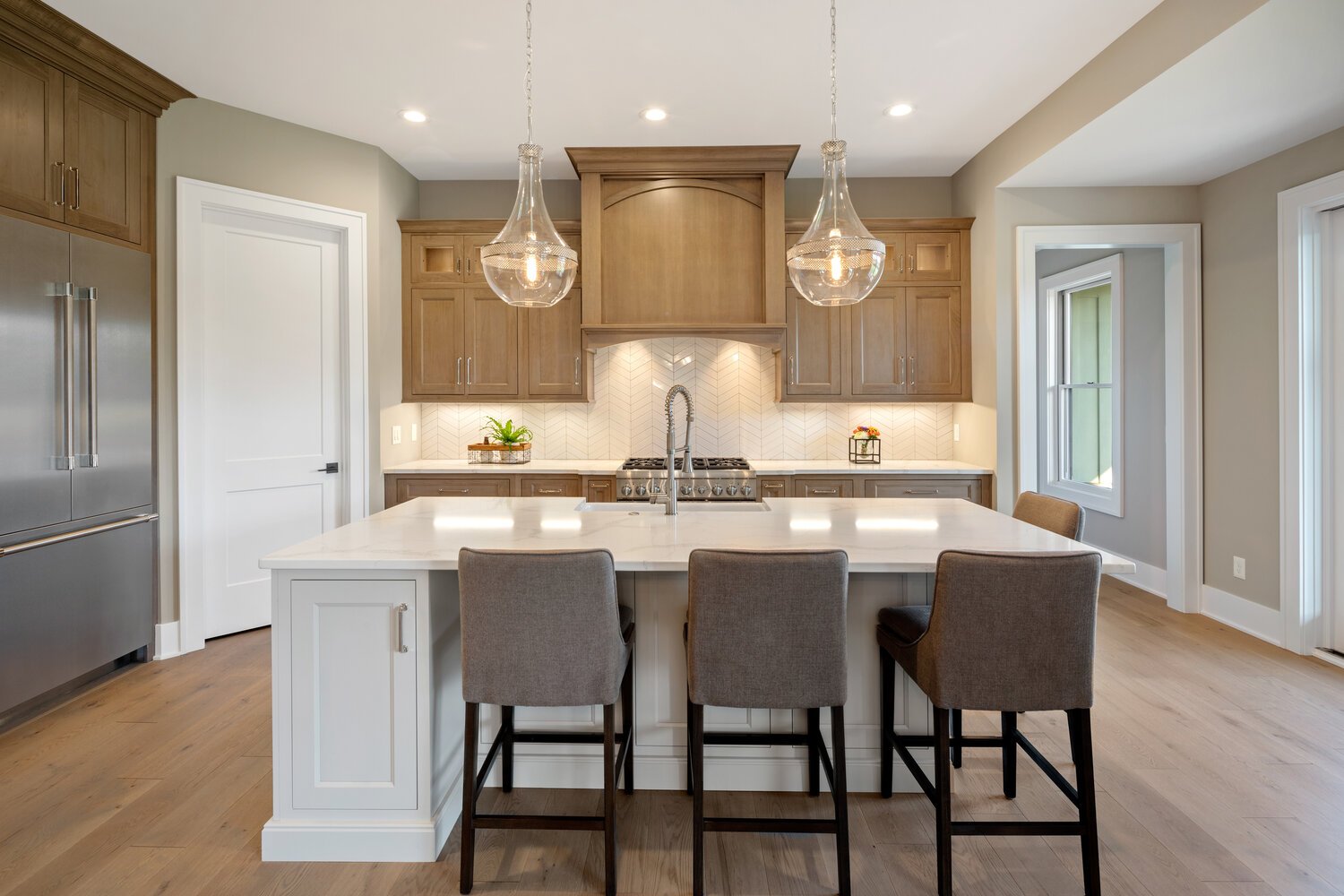 A traditional kitchen with warm wood cabinets, a large island with gray barstools, a tiled backsplash, and stainless steel appliances.