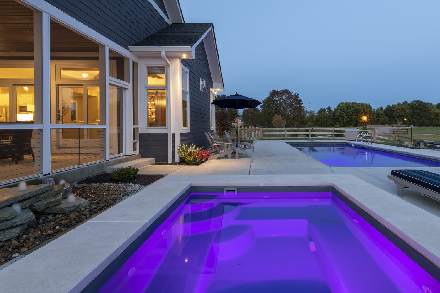 Exterior evening view of a custom home with a deck overlooking a backyard oasis featuring a pool, hot tub, and landscaped surroundings.