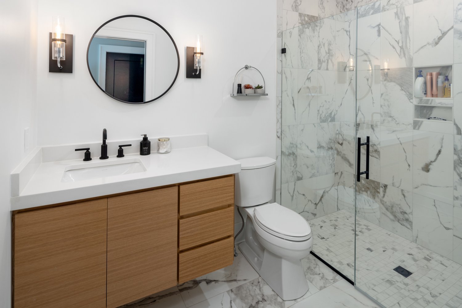 Bathroom with marble tile walls, large round mirror, wooden vanity with white countertop, and glass shower enclosure.