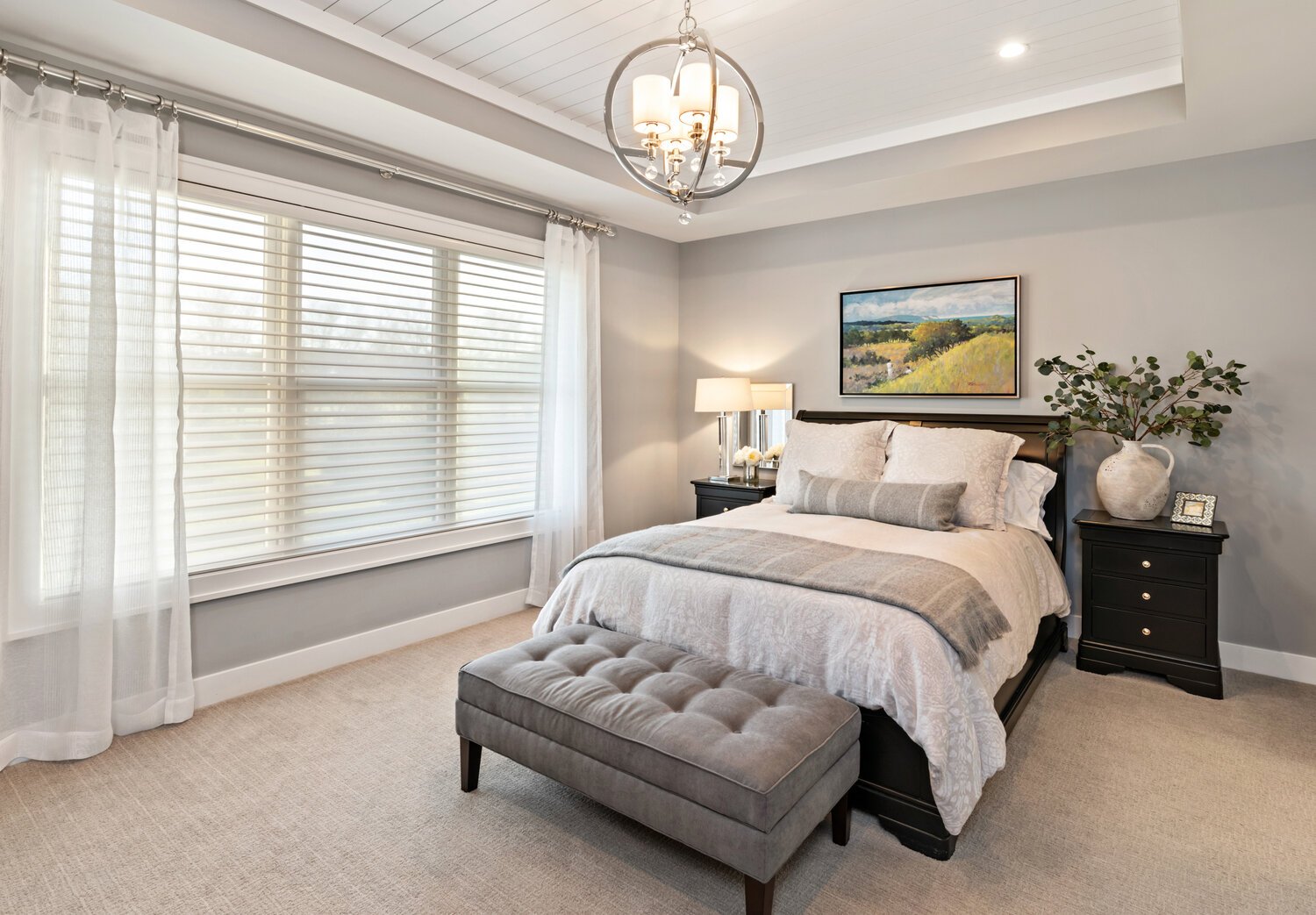 A cozy bedroom featuring elegant neutral tones, plush bedding, large windows with blinds, and a stylish light fixture in a custom home interior.