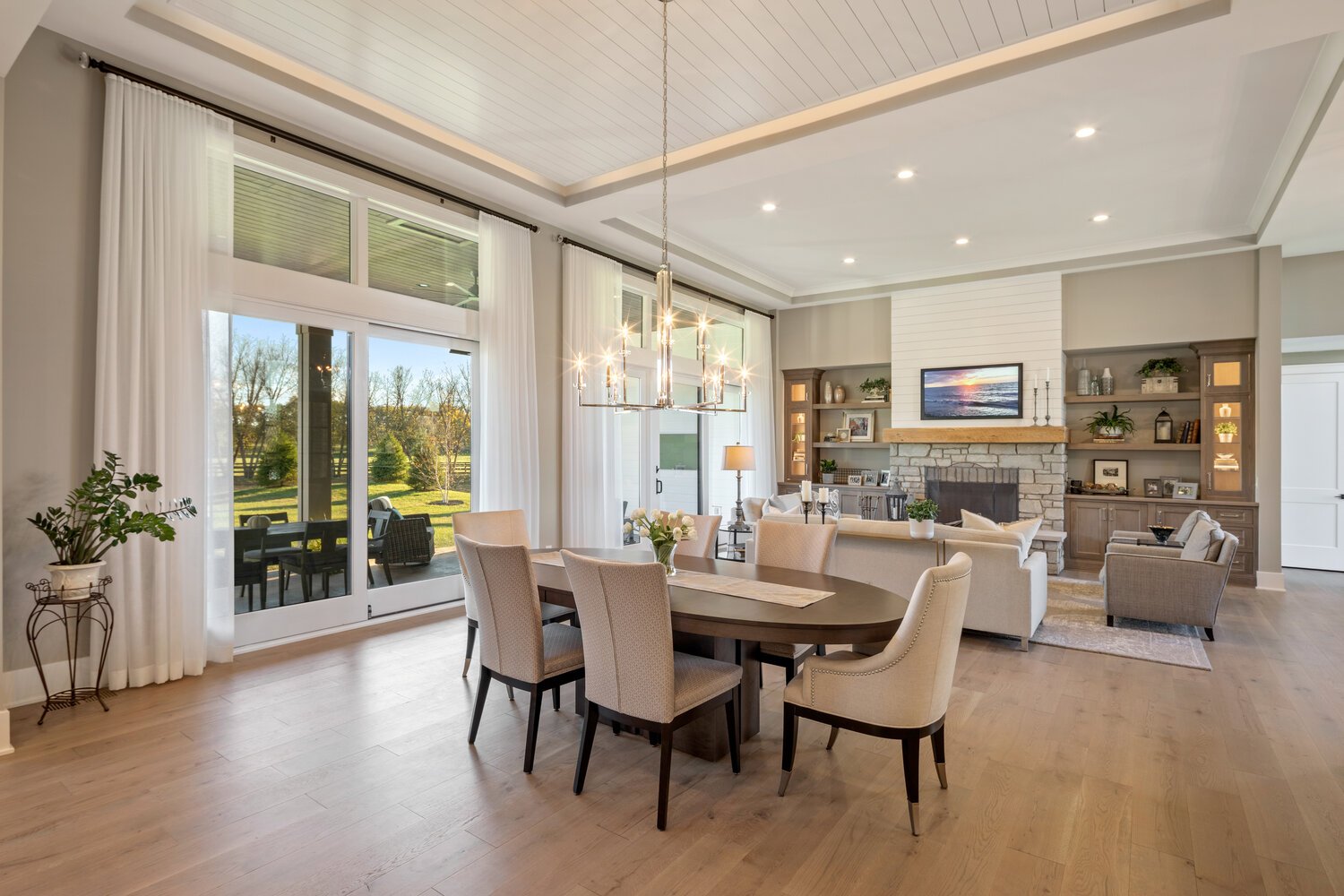 A spacious open-concept living and dining area with vaulted shiplap ceiling, hardwood floors, stone fireplace, and scenic views through large windows.