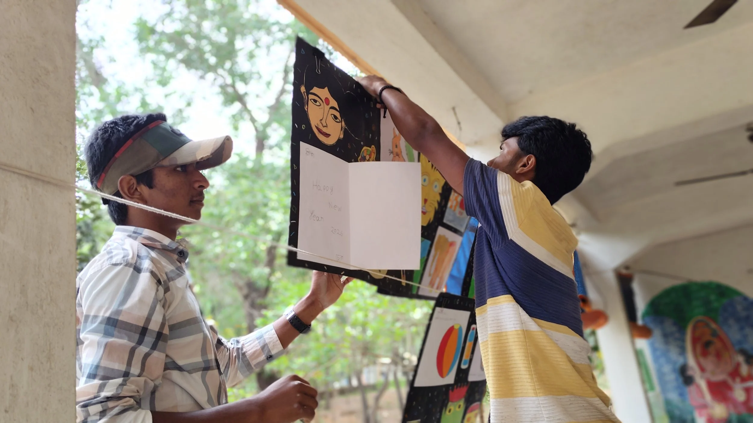 Two men hanging artwork display at an outdoor event, with one holding the artwork and the other making adjustments, with trees and artwork visible in the background.