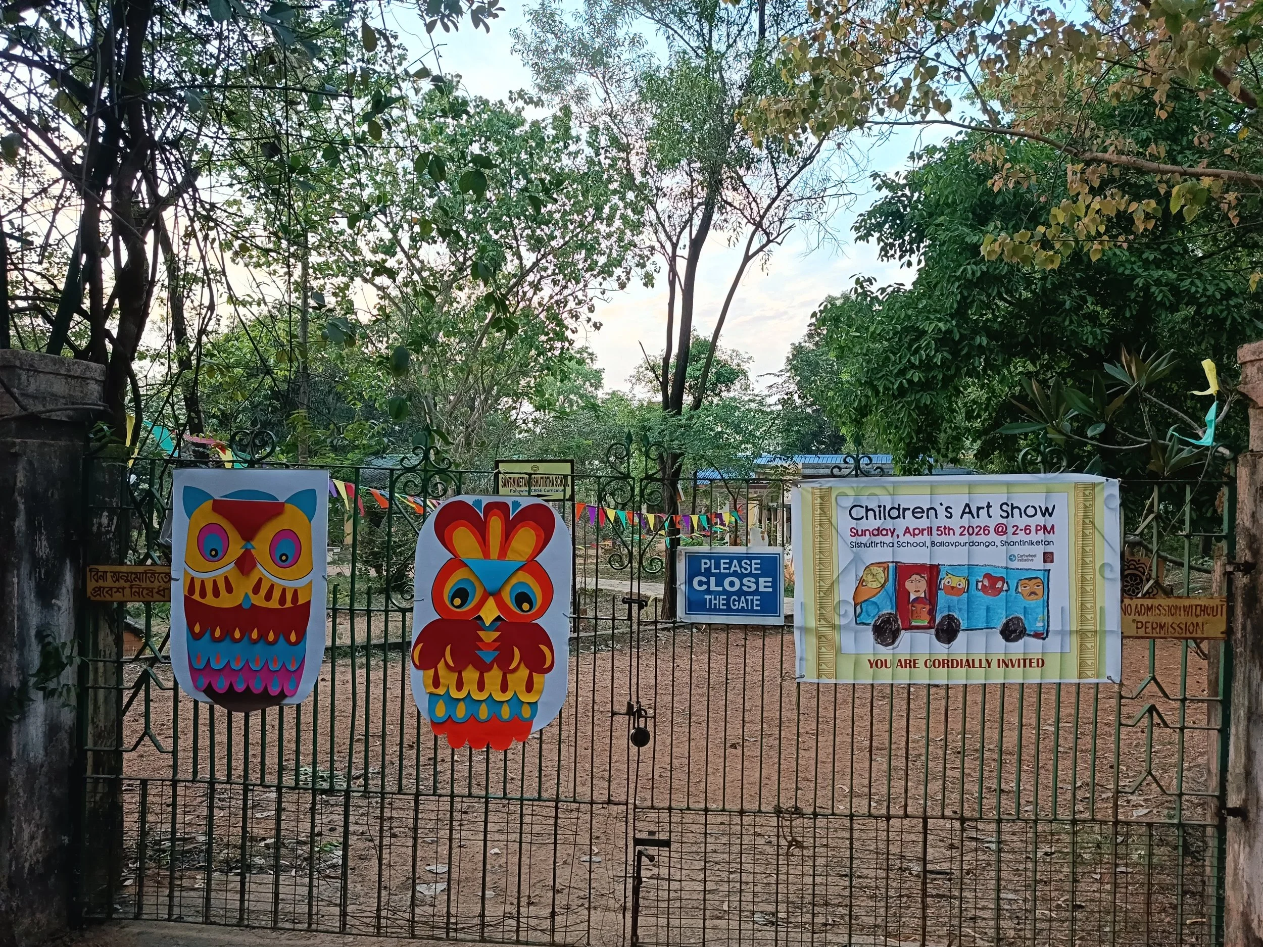 Gated entrance decorated with colorful owl-themed artwork, banners, and a sign for a children's art show at Sishtuthira School, with trees and a cloudy sky in the background.