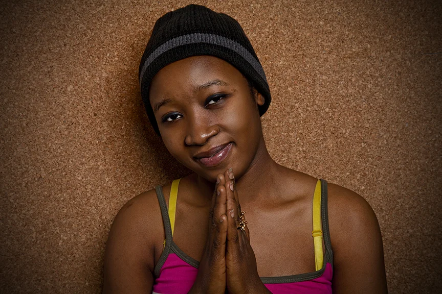 A young woman with a gentle smile, wearing a black beanie and a colorful tank top, standing against a textured brown background.