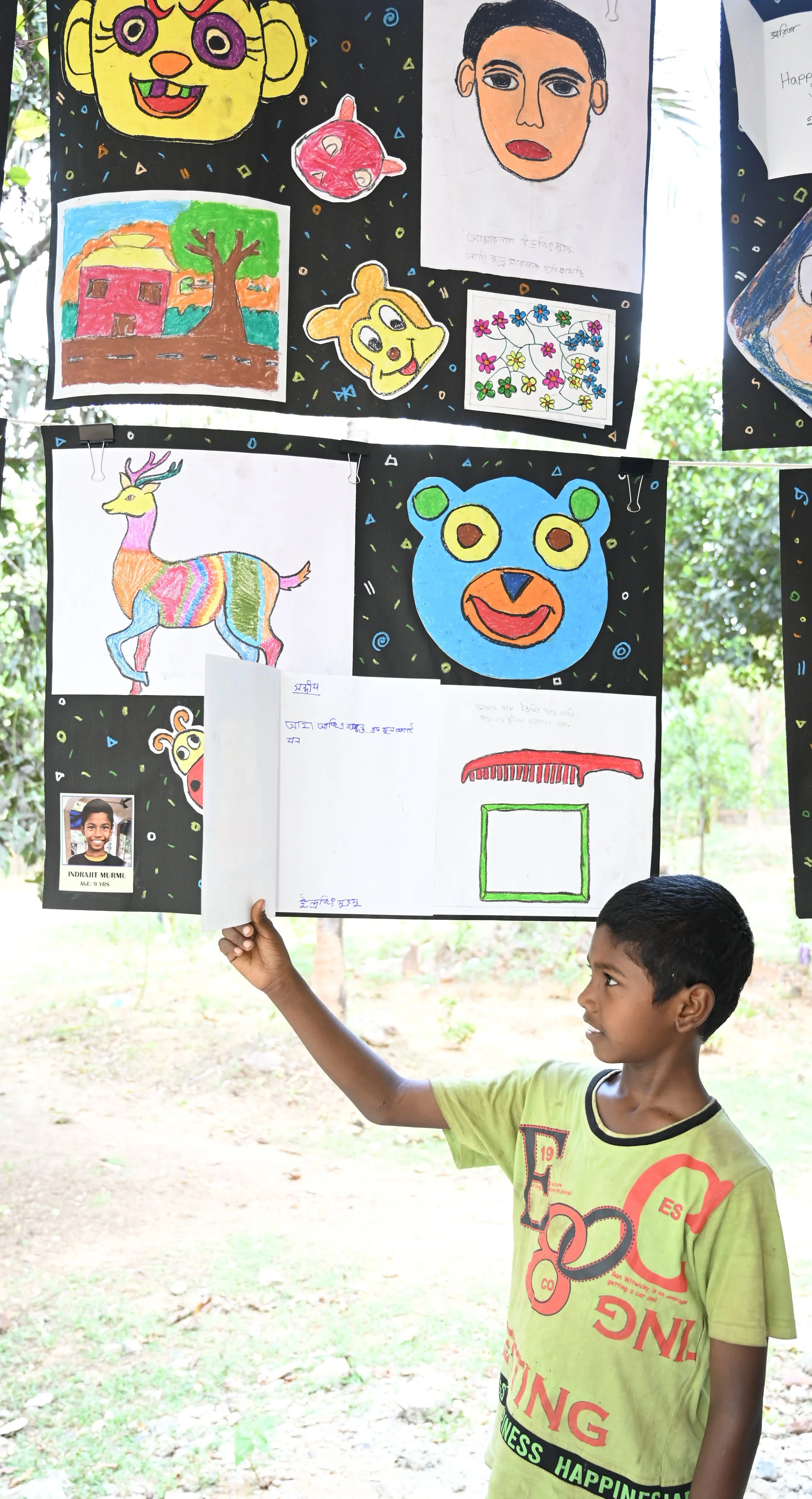 A young boy holding a white paper with handwritten text in front of a display of colorful children's drawings and paintings on a black background decorated with small, colorful confetti-like shapes outdoors with trees and grass visible in the backgro