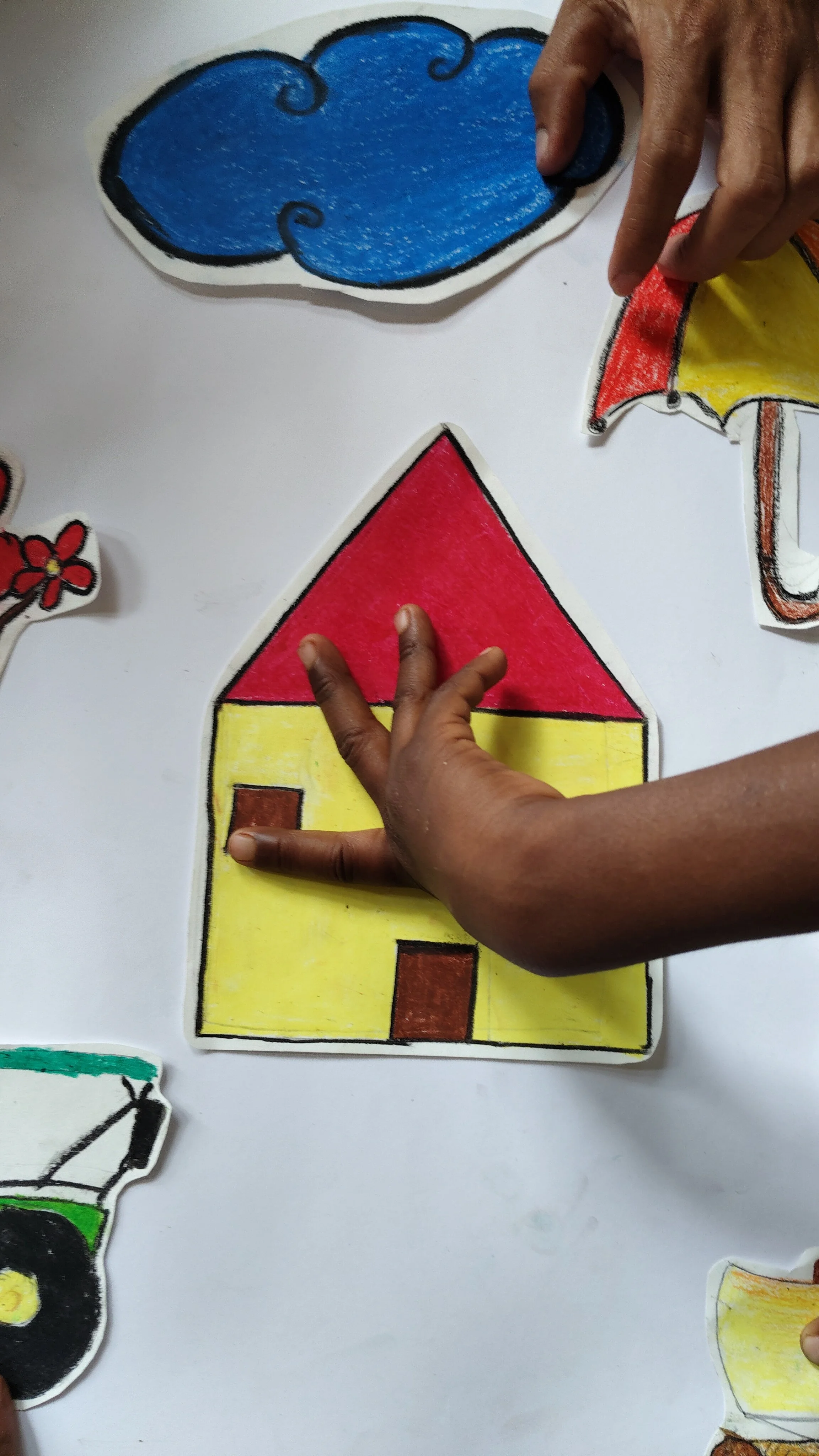 Children's hands interacting with a colorful paper house drawing on a white surface, with other paper drawings partially visible.