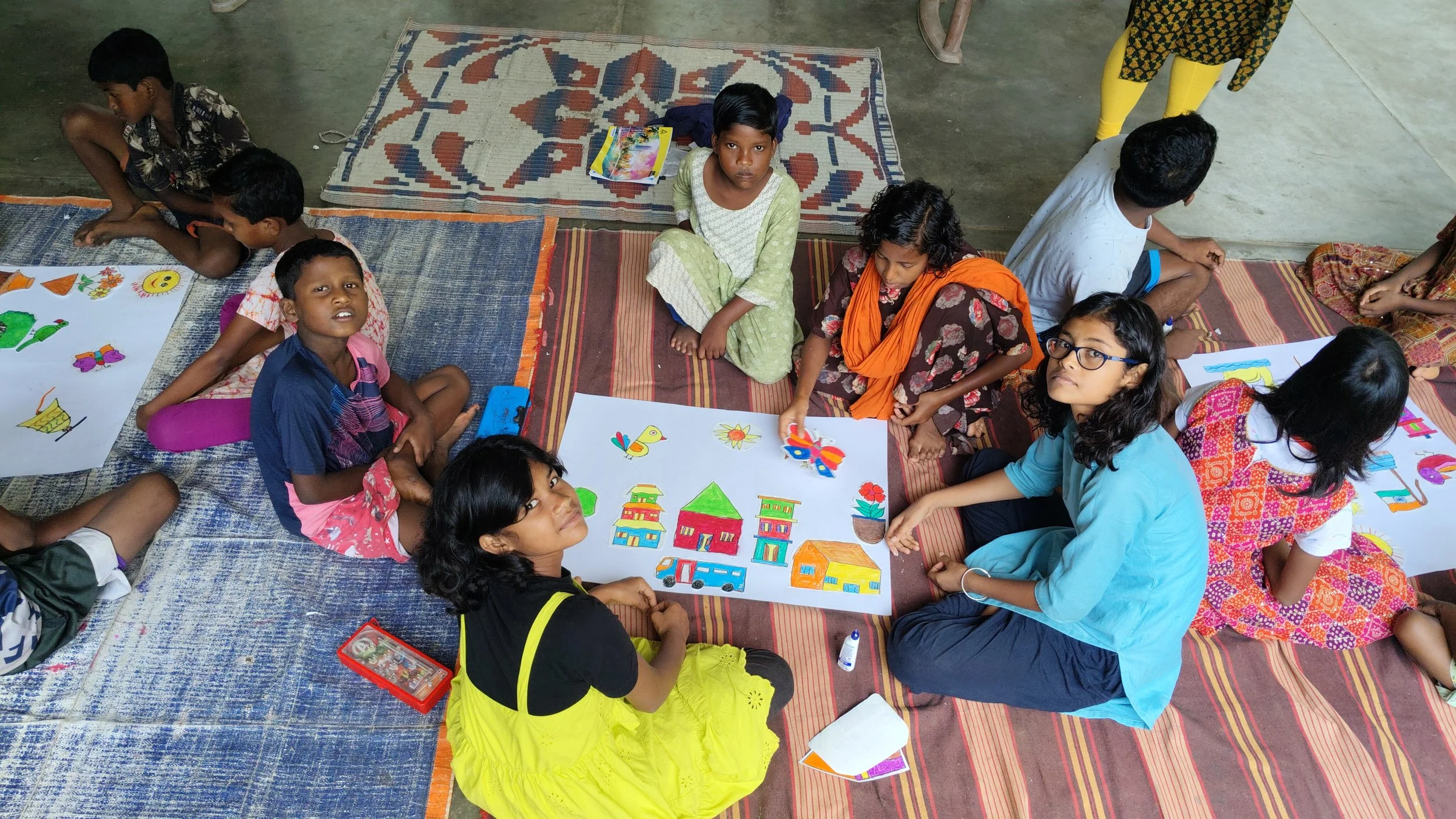 A group of children sitting on a mat on the floor, gathered around colorful drawings and cut-outs of buildings, vehicles, and other shapes, engaged in an art activity.