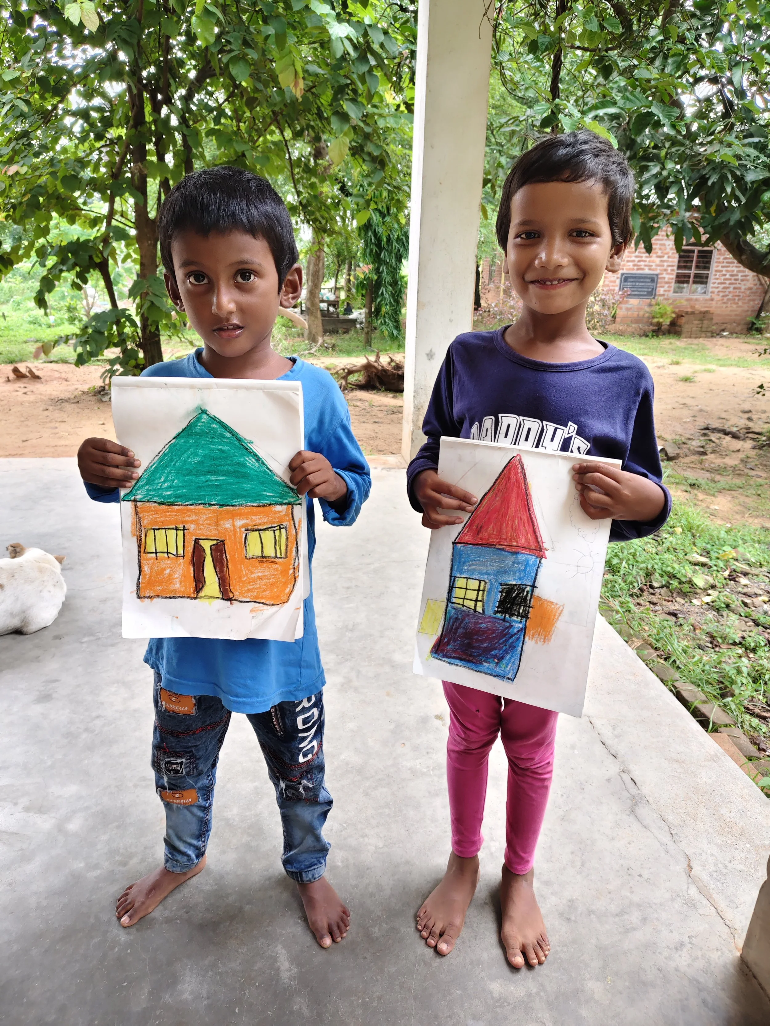 Two children standing outside holding colorful drawings of houses. The boy on the left is wearing a blue shirt and jeans, while the girl on the right is in a dark blue shirt and pink pants. Both children are barefoot. There are trees and a brick buil