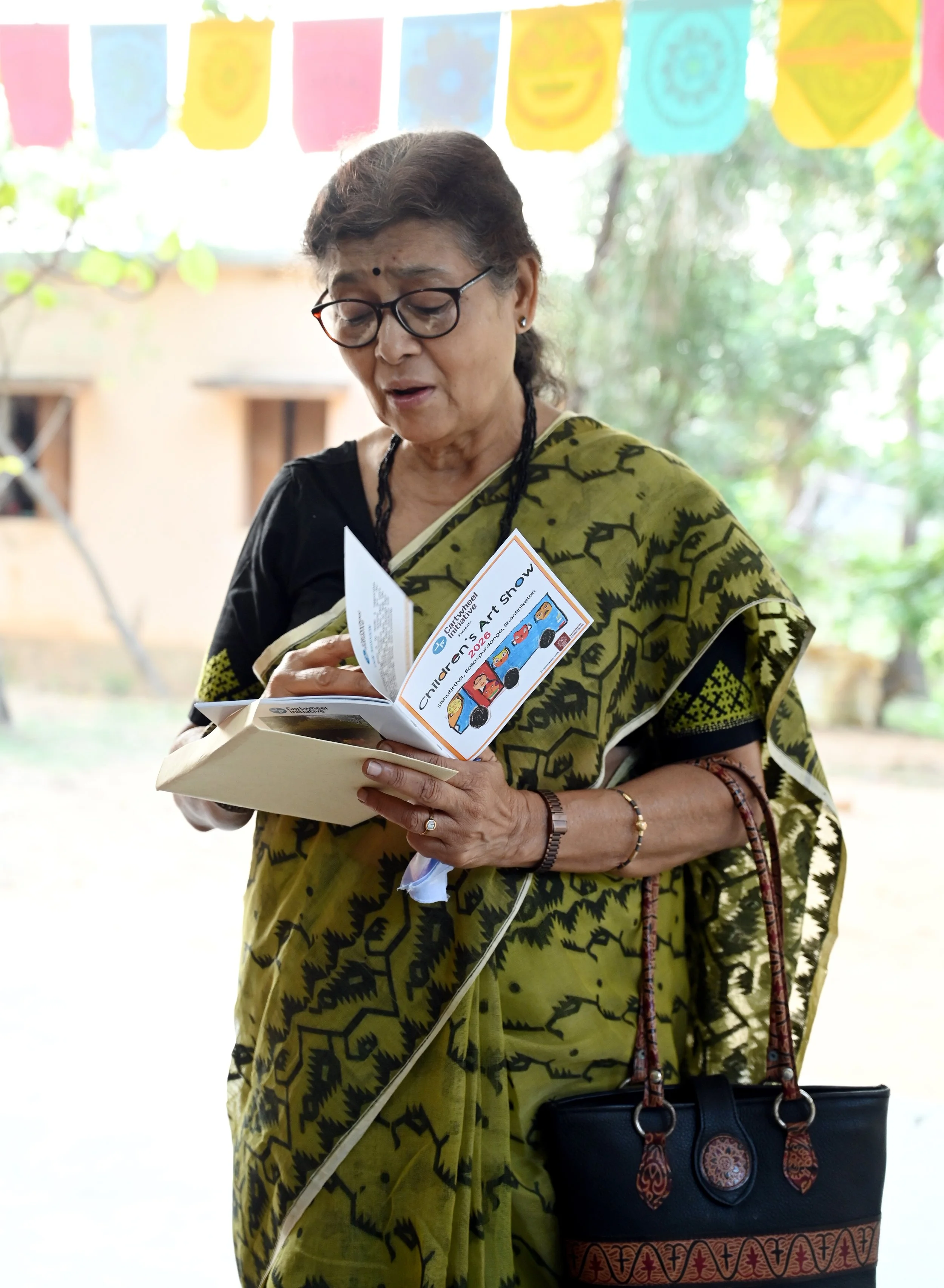 An elderly woman with glasses and a black bindi on her forehead, wearing a yellow-green patterned saree, holds a brochure and a book, standing outdoors with trees in the background, behind a colorful banner and next to a black handbag with orange and