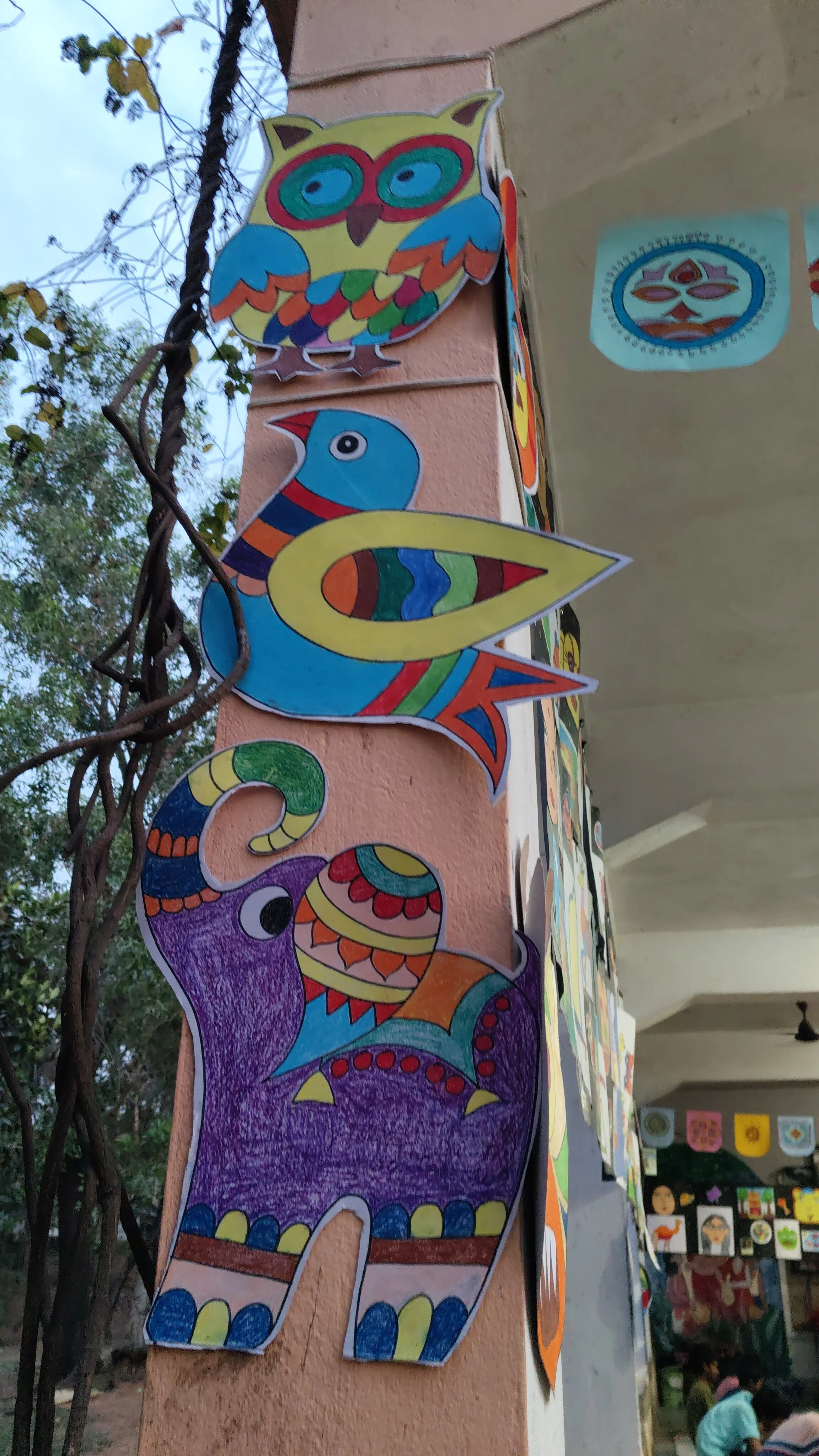 Colorful paper cutouts of various animals, including an owl and a bird, are attached to a corner of a building's wall, outside, with trees and part of a marketplace in the background.
