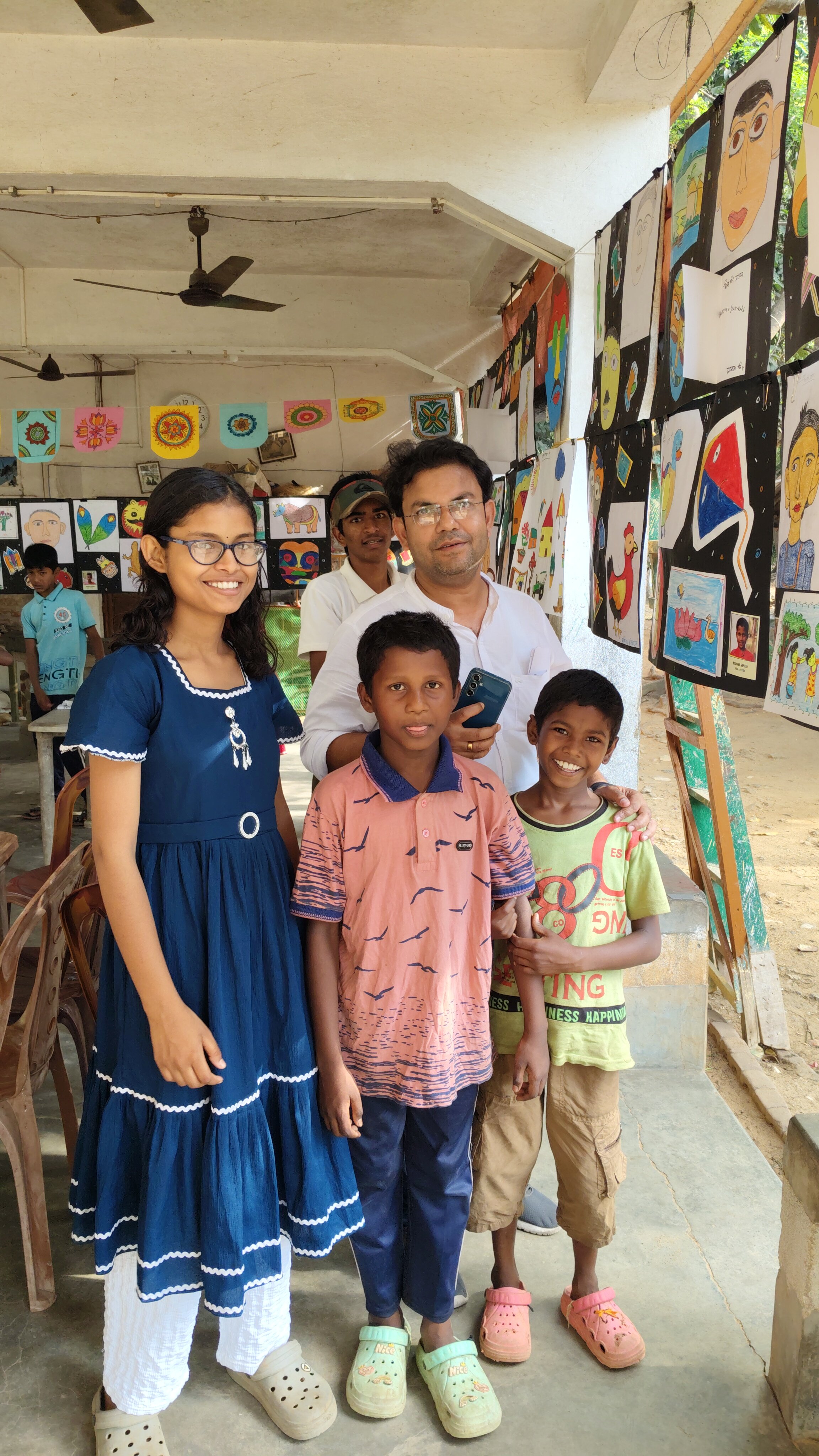 A group of five people, three children and two adults, standing indoors in front of a display of colorful artwork hanging on the walls. The children are smiling, and one of the adults is holding a smartphone.