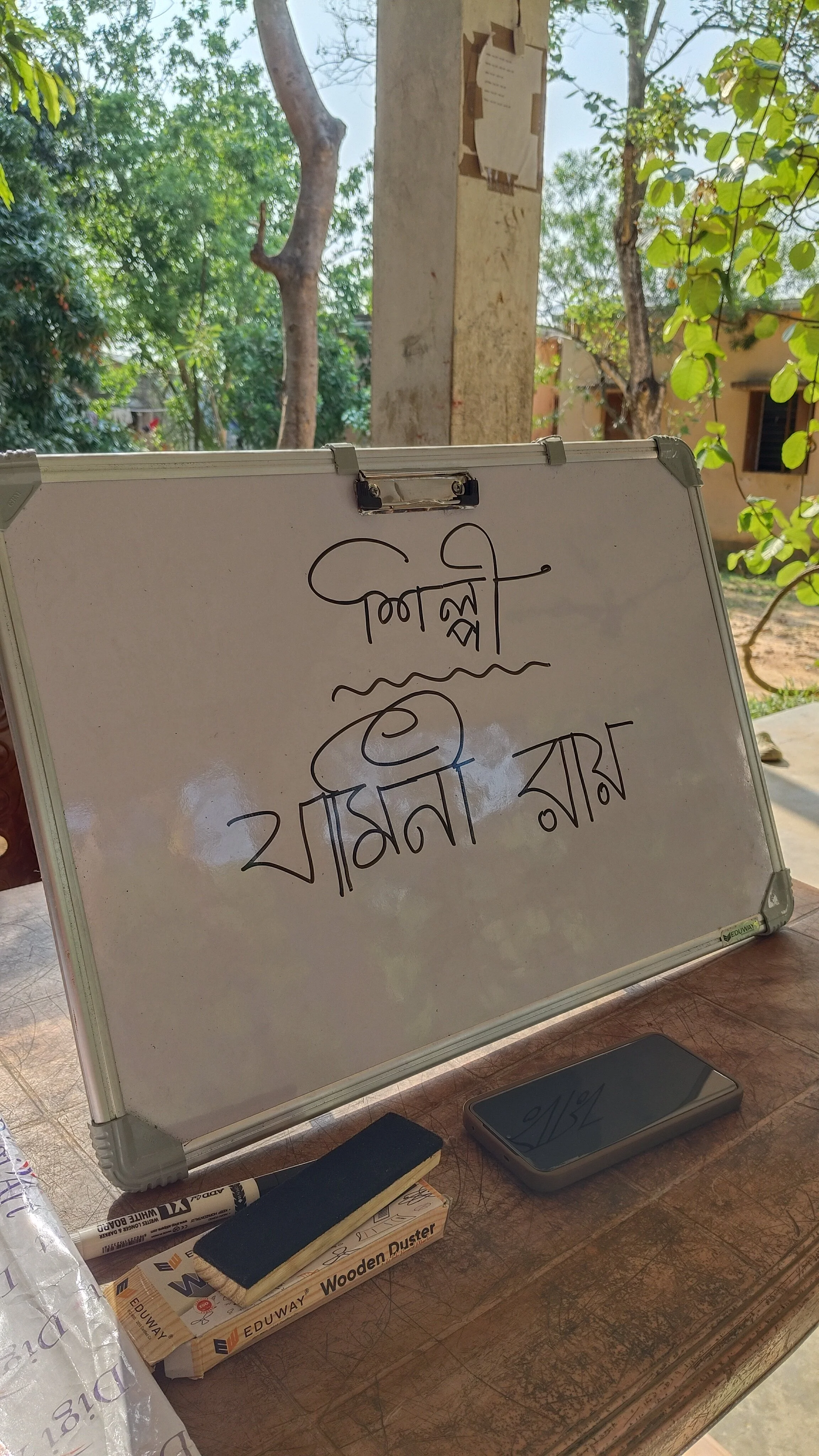 Whiteboard with handwritten text in Bengali script, placed on a wooden table outdoors with trees and a building in the background.