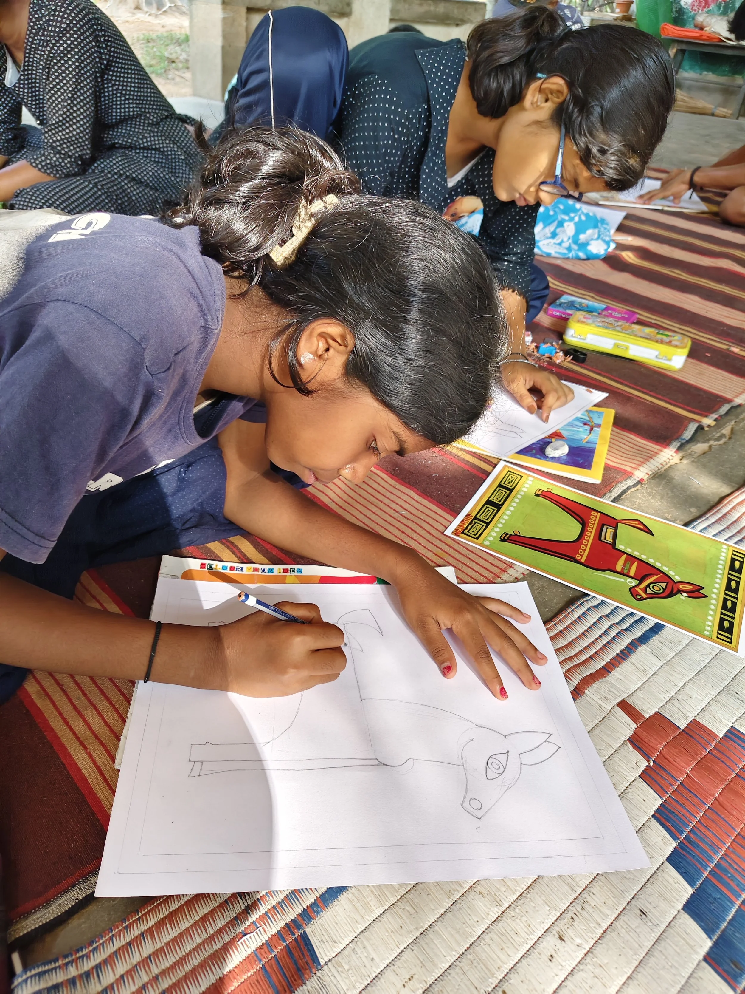 Children are drawing animal pictures on sheets of paper while sitting on a mat outdoors.
