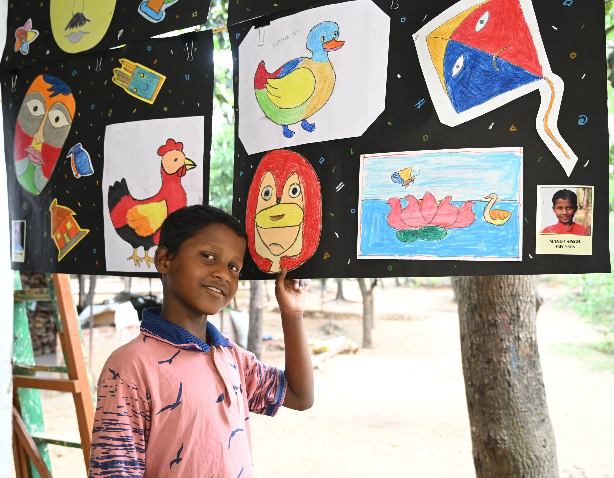 A young boy is smiling and holding up a colorful drawing of various animals and objects, displayed on a black background with small, colorful confetti-like decorations. The boy is outdoors, near trees, and there are other children and a photo of a yo