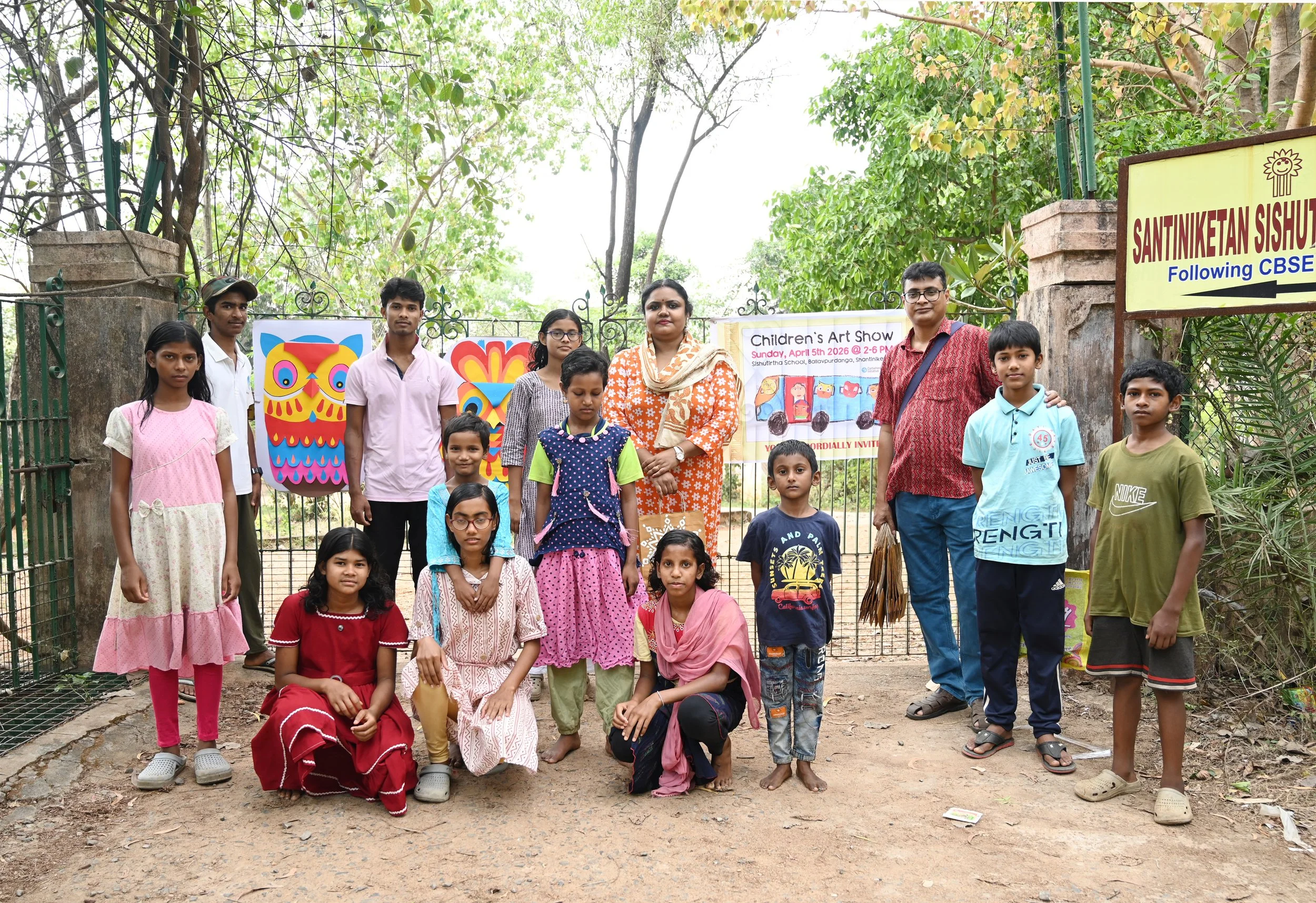 A group of children and adults standing outside in front of a gate with colorful artwork and a banner for a children's art show, surrounded by trees and greenery.