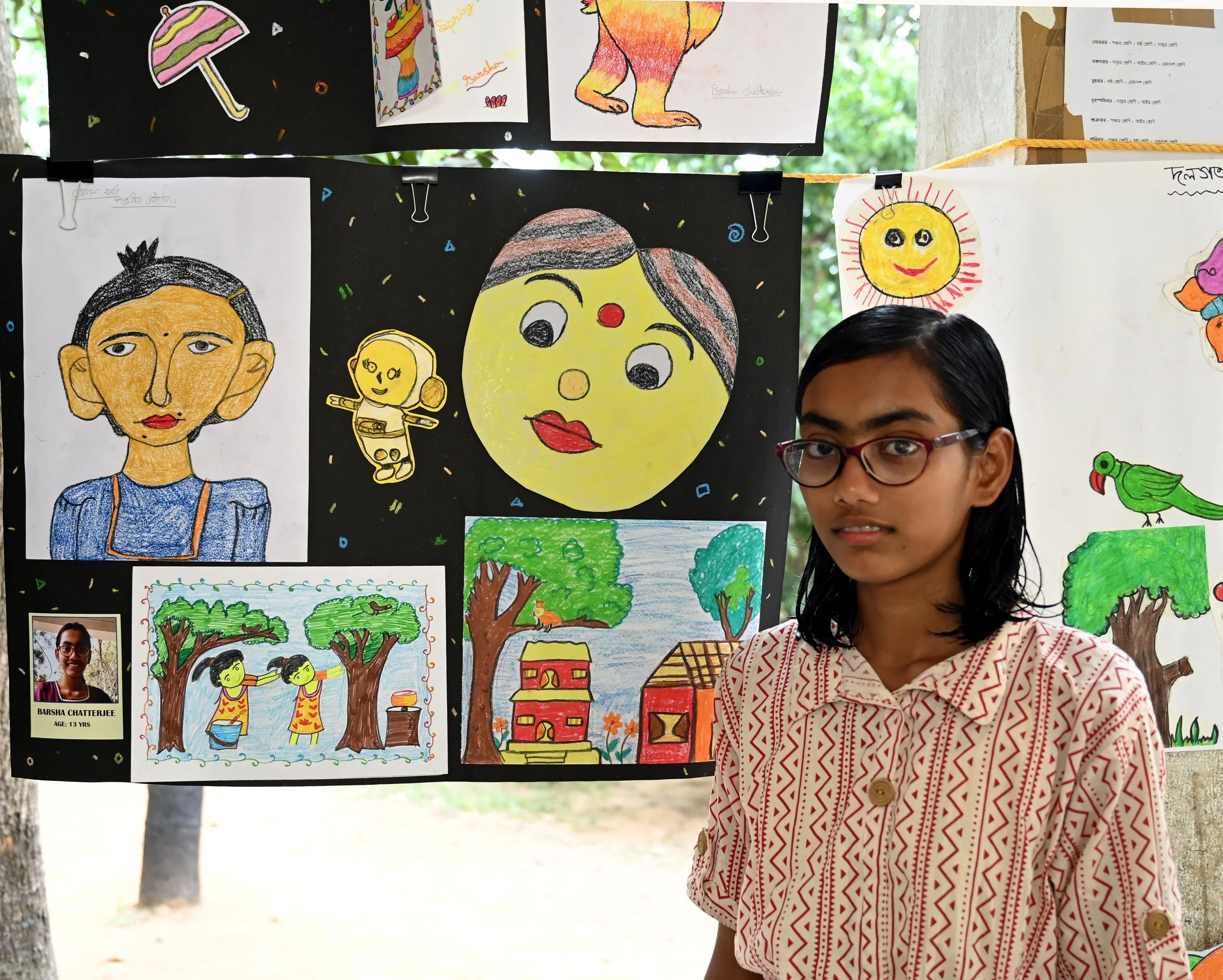 A display of colorful children's artwork hang on a black background. A girl with glasses and striped shirt stands in front of the display. The artwork includes portraits, animals, and nature scenes.