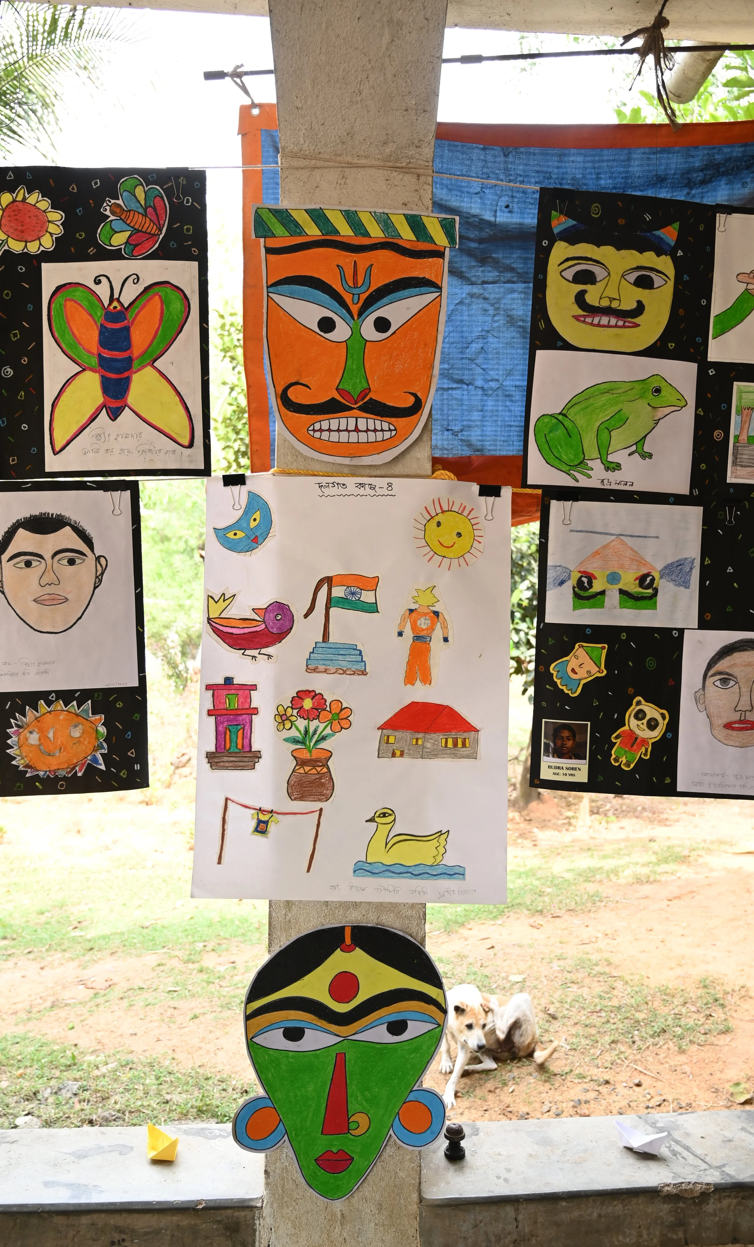 Colorful traditional mask with green face, red nose, blue eyes, orange circles on cheeks, and decorative blue and orange earrings. The mask is hanging in an outdoor setting with other artistic drawings and masks displayed around it, and a dog lying o