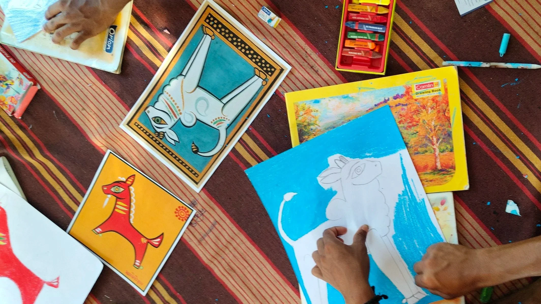 Child's hands sketching a horse on blue paper surrounded by coloring books and crayons on a striped tablecloth.