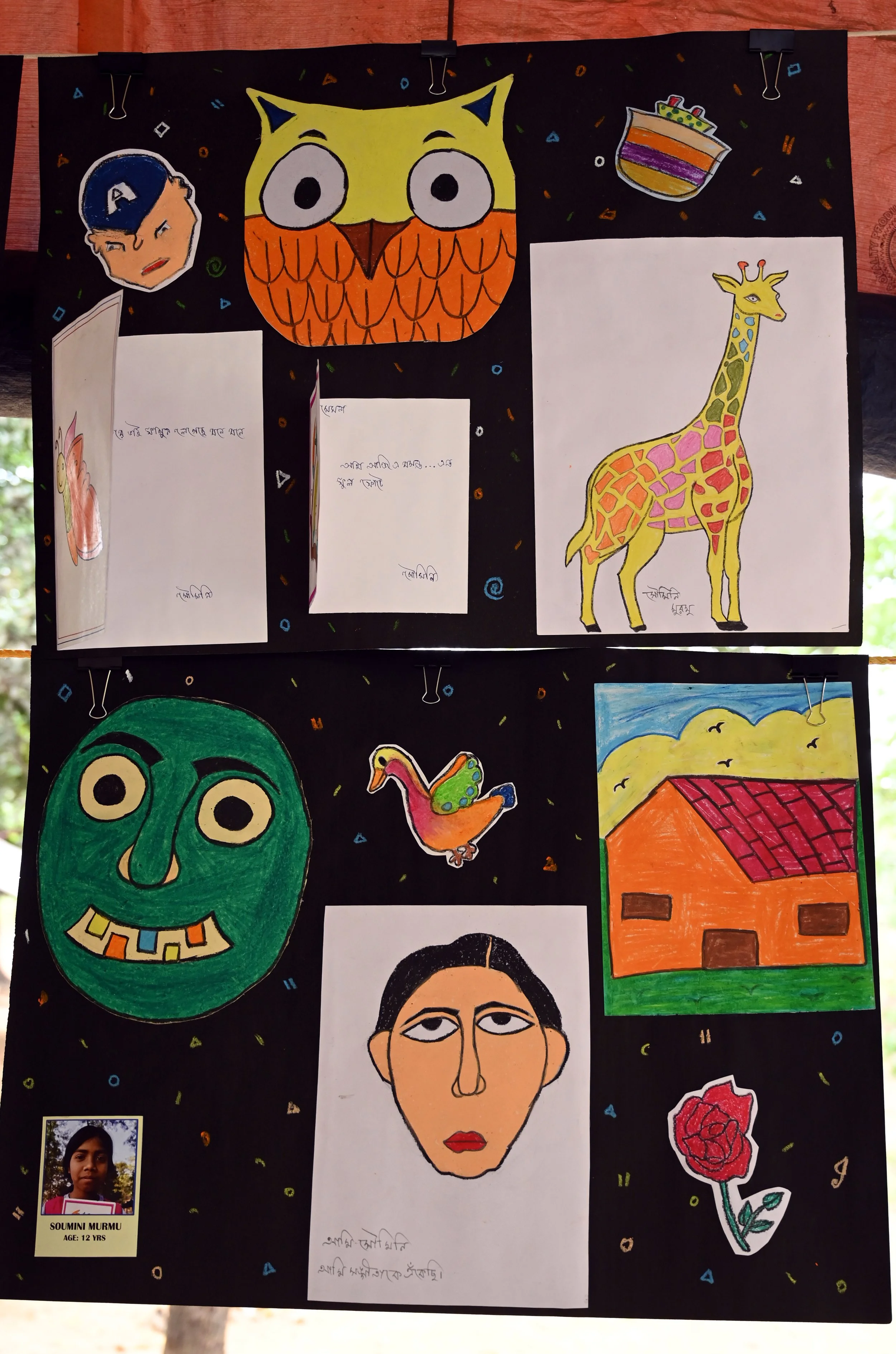 Colorful drawings and cutouts of various objects and faces pinned onto a black background, including a cartoon face, a giraffe, a house, a mask, and a flower, with some handwritten notes.