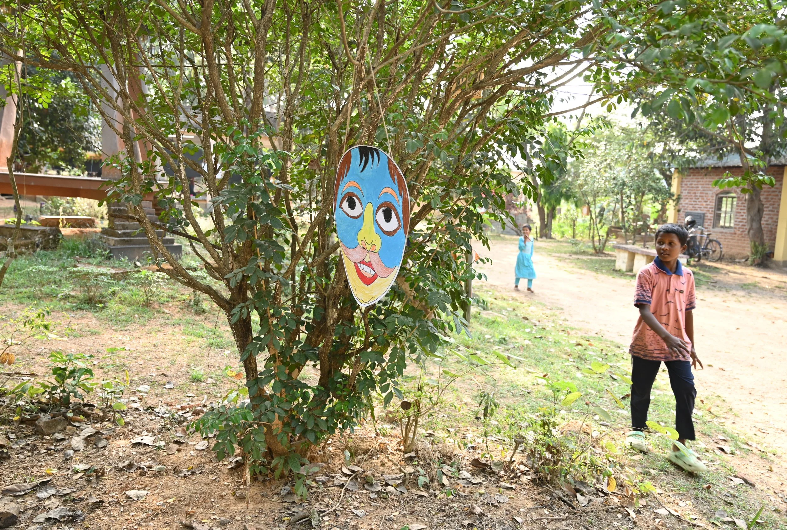 Children playing outdoors near a tree decorated with a colorful painted face mask. The setting appears to be a rural or village area with houses in the background and children walking around.