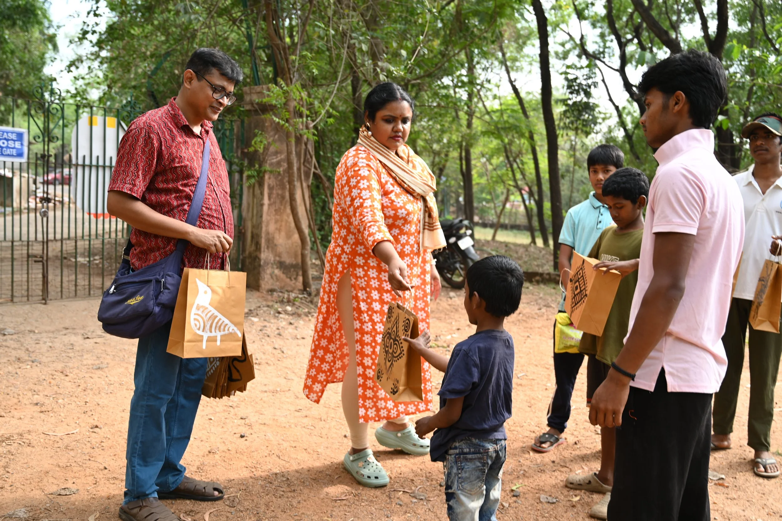 A group of people, including children and adults, are standing outdoors in a park with trees and a fence in the background. They are holding brown paper bags and appear to be participating in an outdoor event or activity.