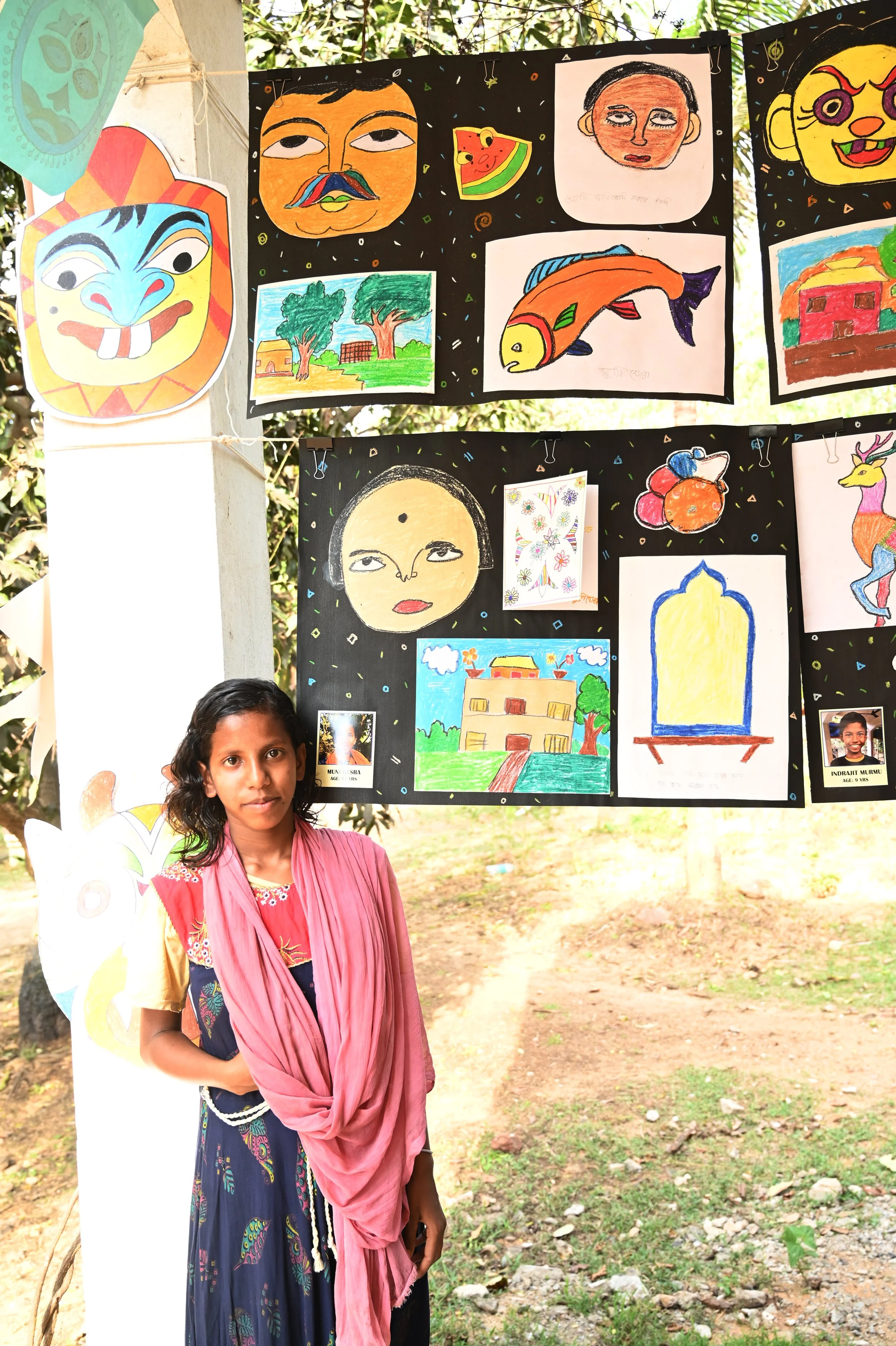 A young girl stands outdoors in front of a display of colorful children's drawings and paintings hanging on a black fabric background. The artworks include portraits, a fish, a house, a tree, and abstract designs. The girl wears a pink scarf over a t
