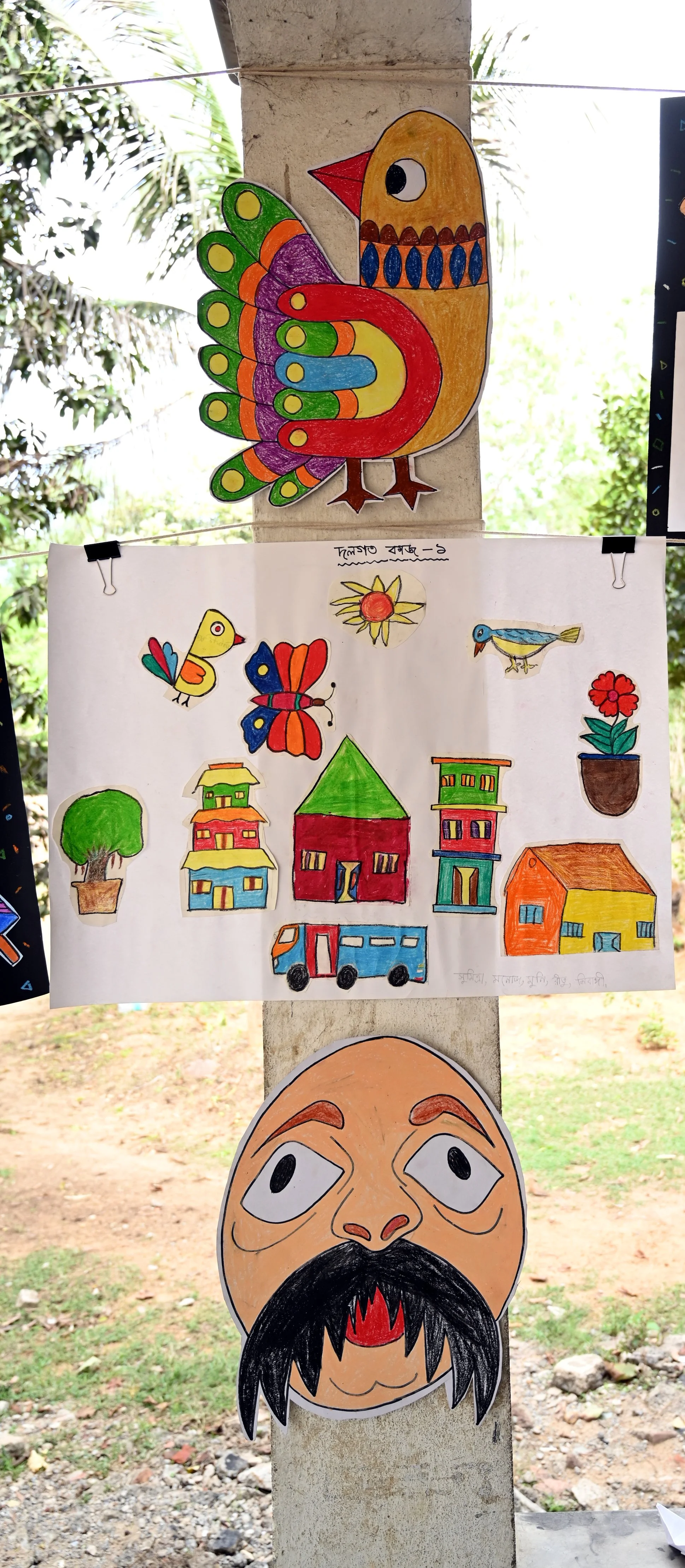 Colorful children's drawings of a bird, flowers, a house, a tree, cars, and other nature and village scenes displayed outdoors.