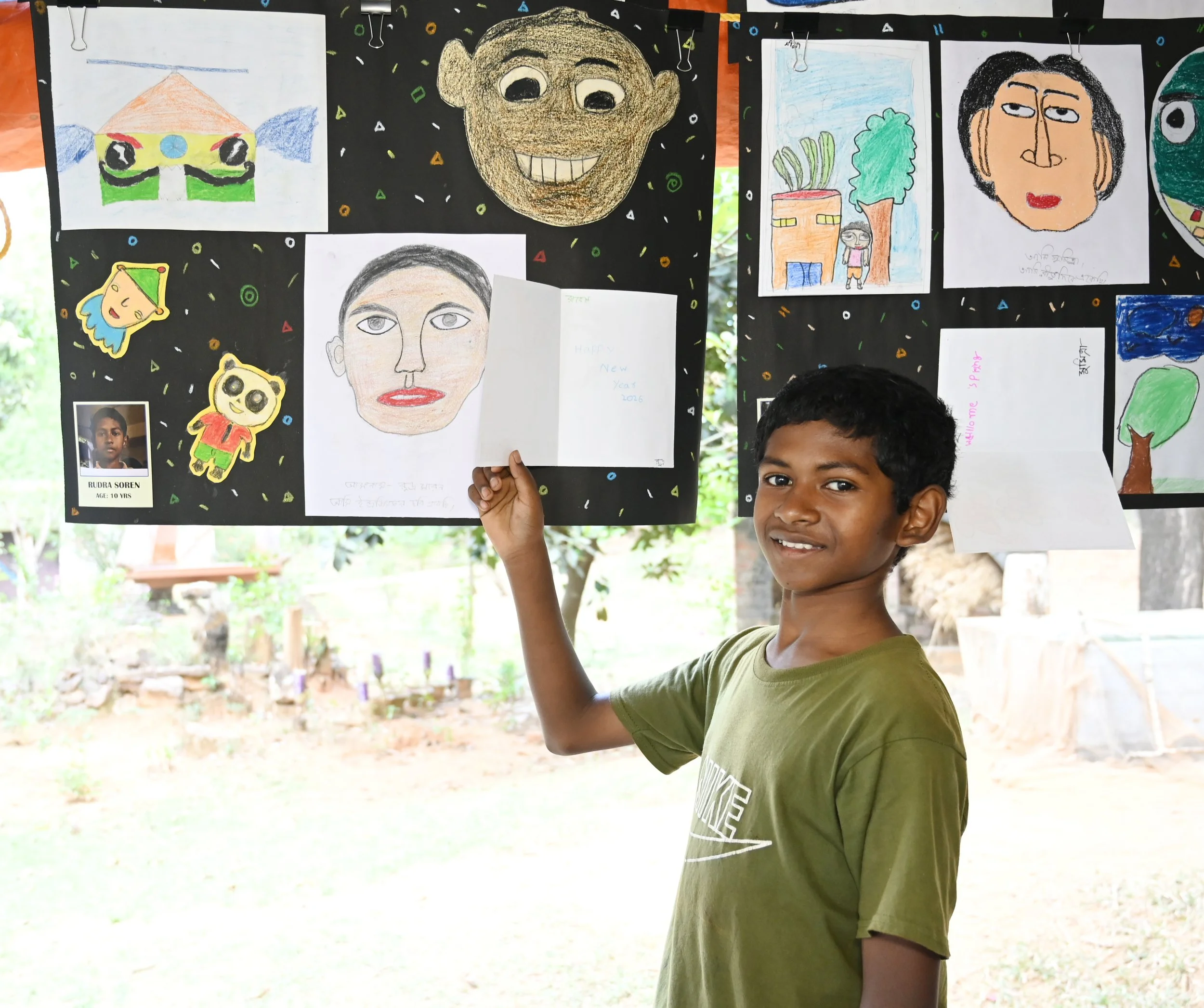 A young boy holding up a drawing at an outdoor art display. The display features various colorful drawings of faces and objects on black background boards, with some small photos and handwritten notes.
