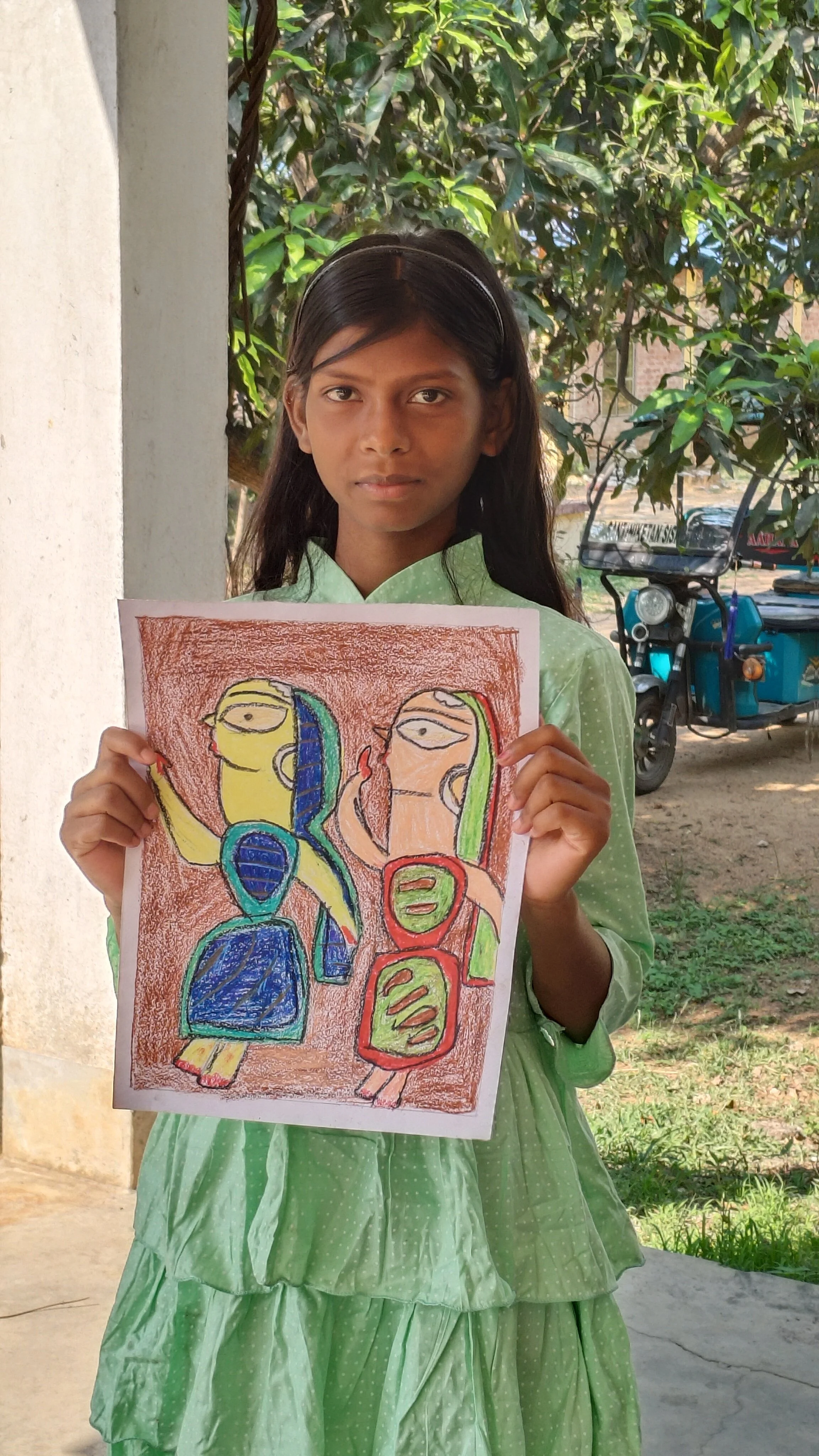 A young girl in a green dress is holding a colorful drawing of two abstract human figures, standing outdoors with trees and a blue vehicle in the background.