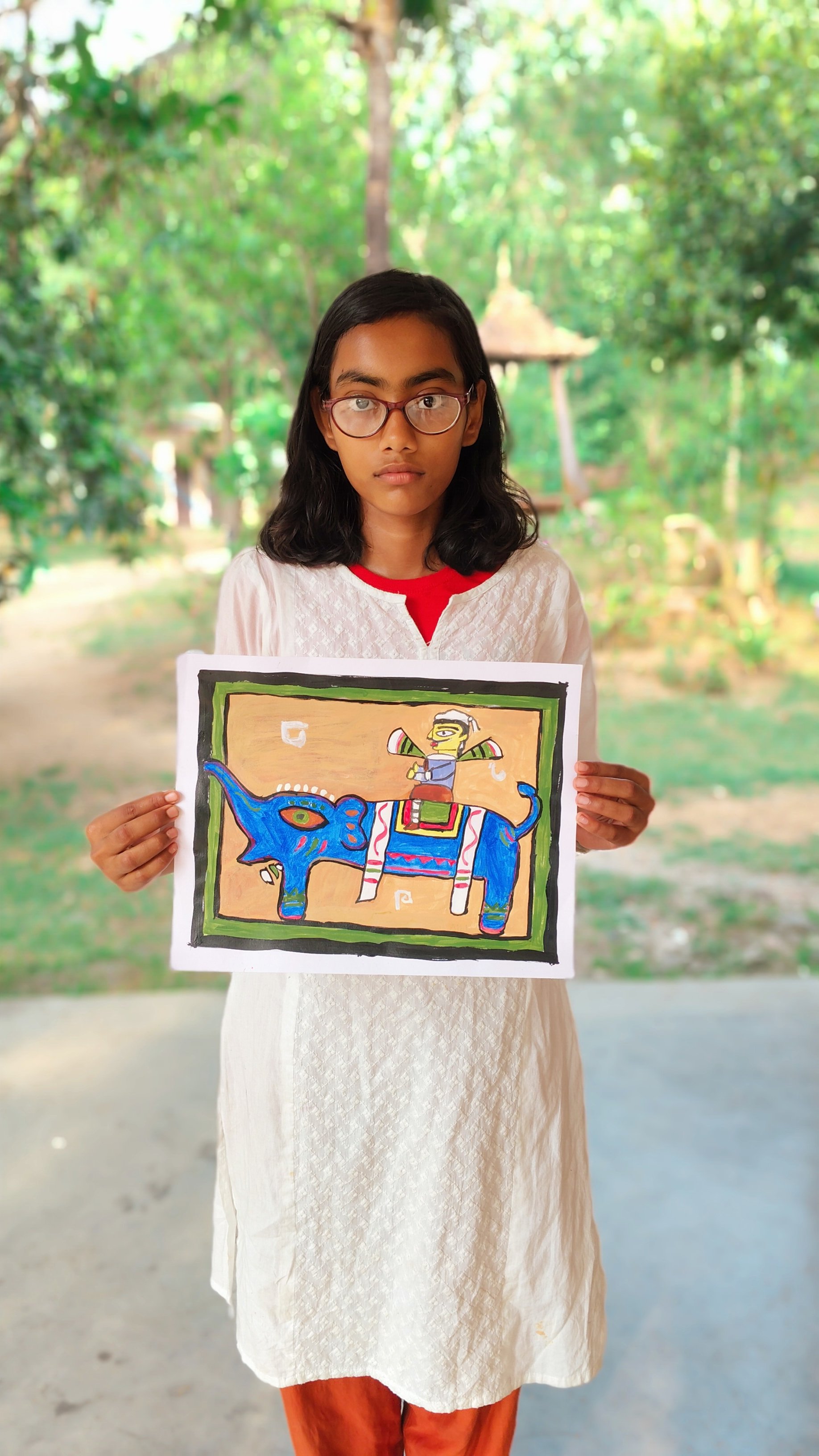 A girl with glasses holding a colorful drawing of a decorated blue elephant with a person riding it, set against a lush, green outdoor background.