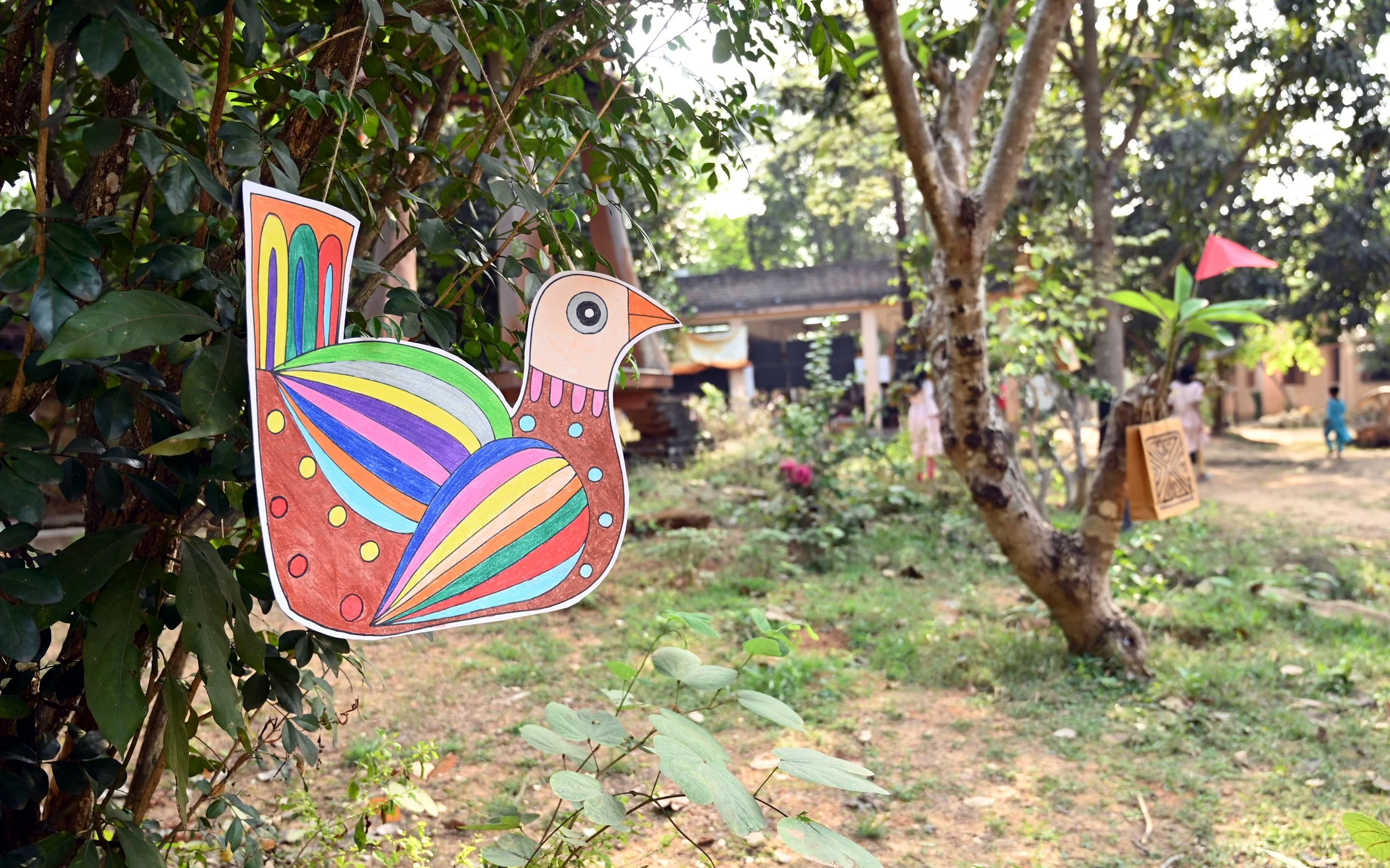 Colorful paper cutout of a stylized bird with rainbow feathers hung on a tree in an outdoor park or garden.