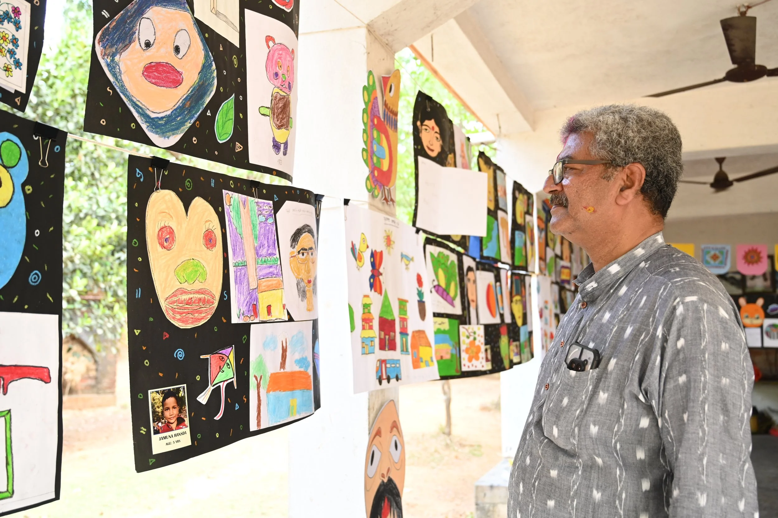 A man with curly gray hair, glasses, and a patterned shirt is looking at children’s artwork displayed on a board at an outdoor art exhibit. The artwork includes colorful drawings and paintings, some depicting faces, animals, and abstract designs.