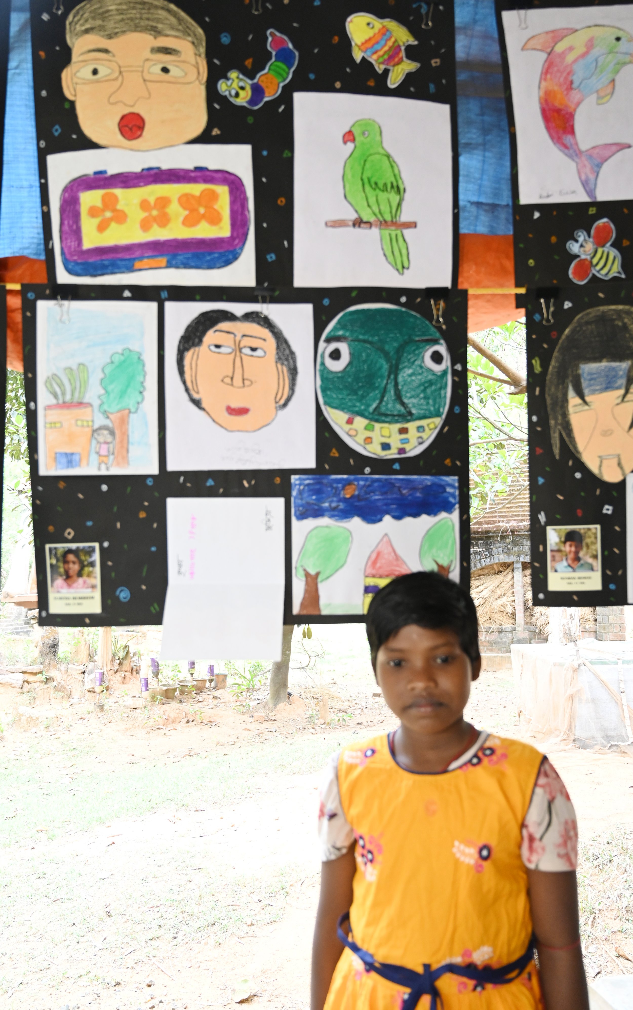Children's artwork is displayed on a black background, featuring colorful paintings of a human face, fish, a parrot, a house with trees, and a butterfly. A boy in a yellow dress with floral patterns stands in the foreground, with trees and a wooden s