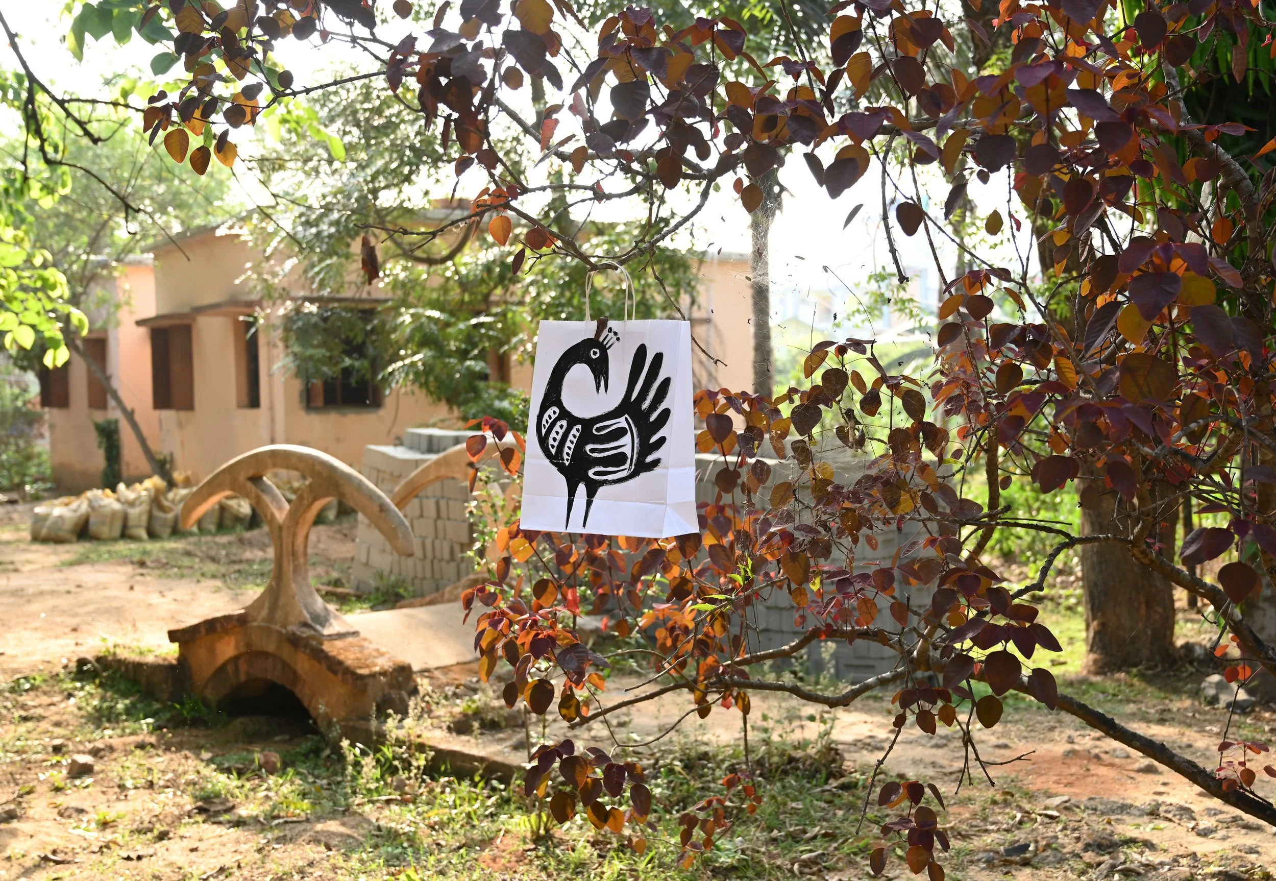 A garden scene with flowering tree branches, a hanging artwork with a black bird illustration, trees, a building in the background, and decorative wooden log and brick structures on the ground.