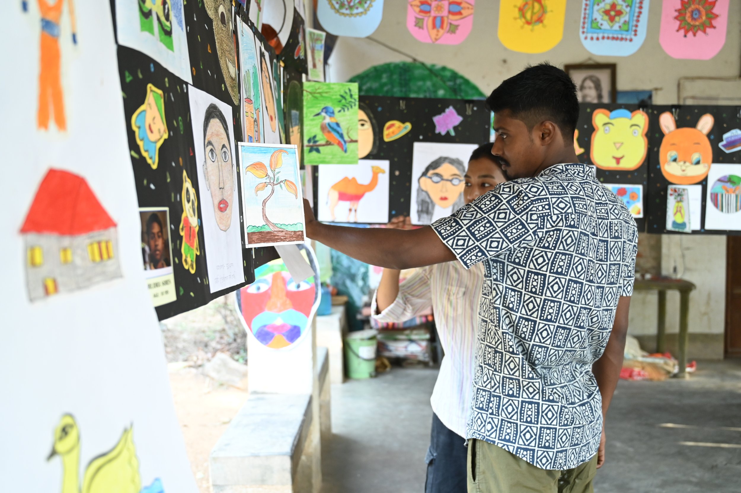 Two young men looking at colorful artwork displayed on a white and black board in an open classroom or gallery space. The background shows various vibrant art pieces and decorations hanging on the walls.