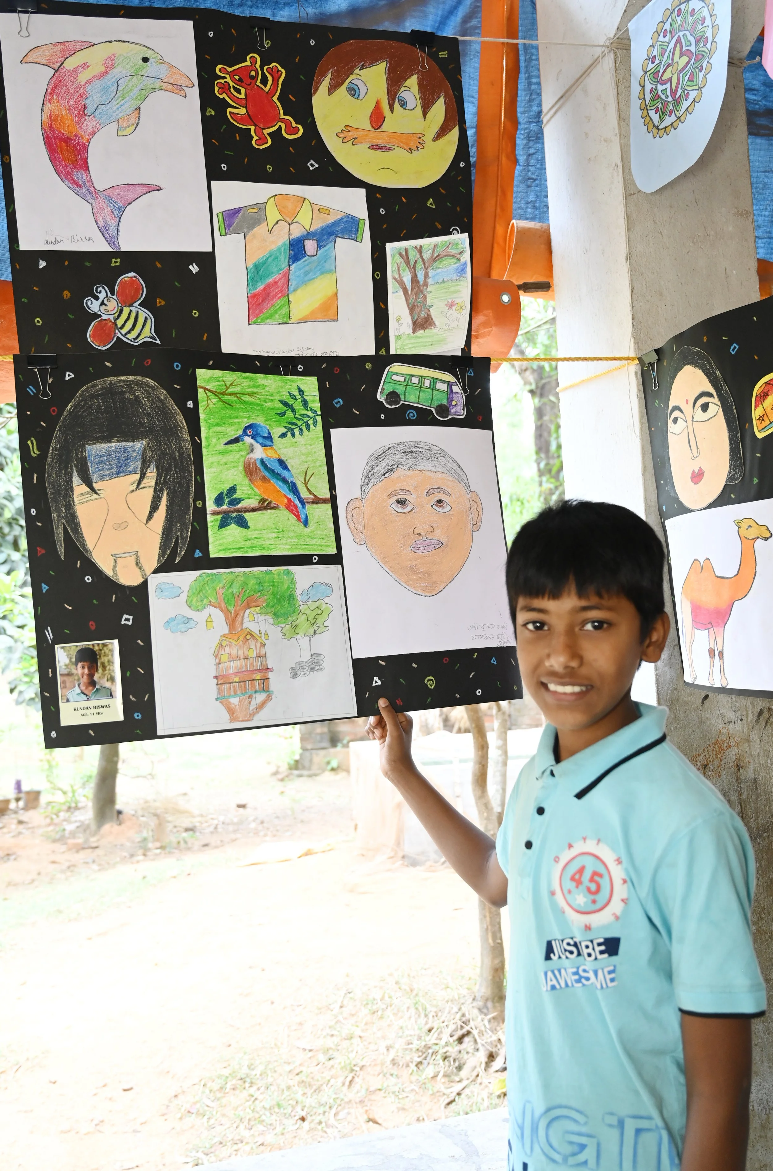 A boy in a light blue shirt smiling and holding a black display board with children's drawings, including a dolphin, a butterfly, a bird, a tree house, and human faces. The outdoor setting has trees and a sandy ground in the background.