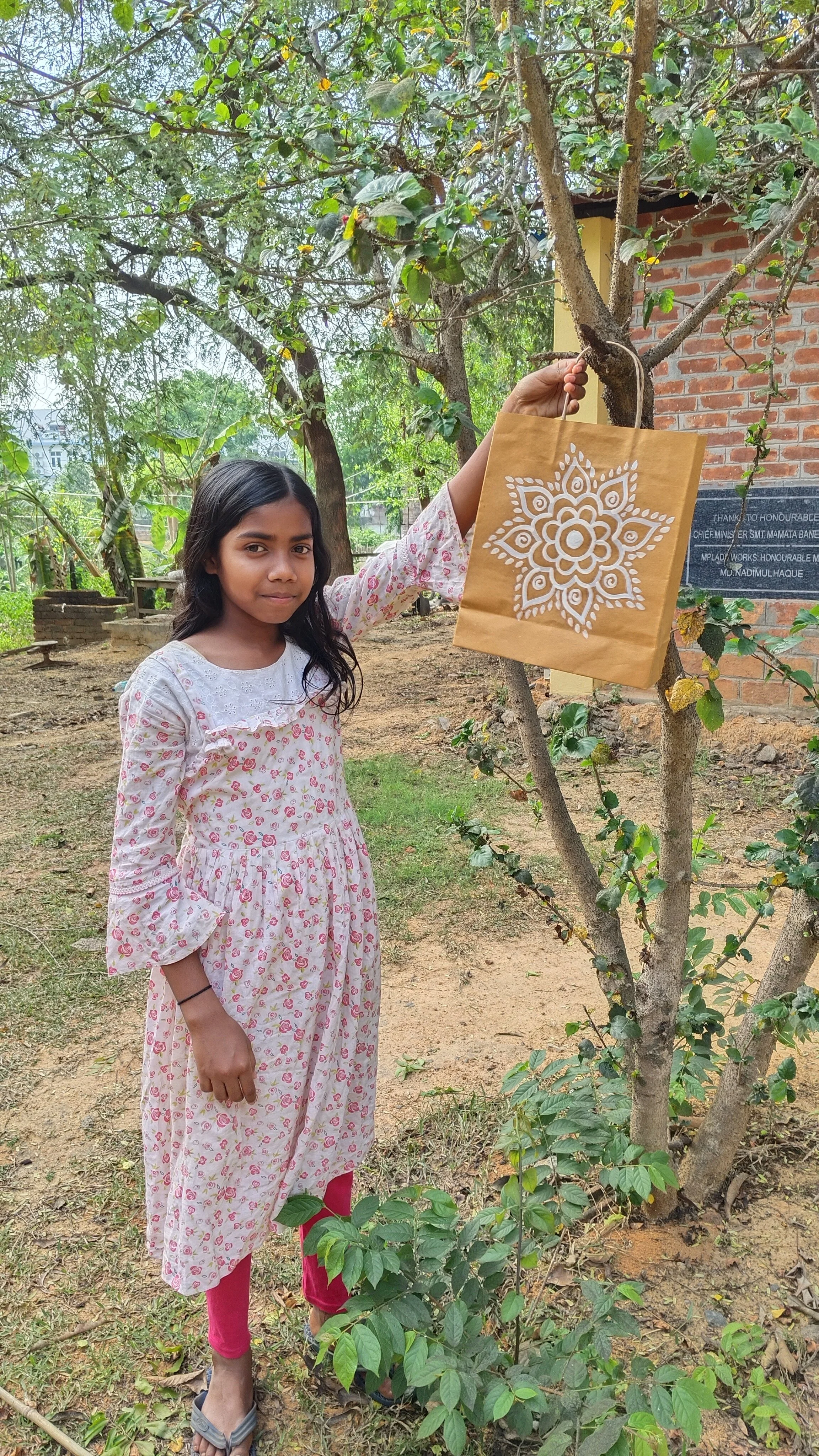 A young girl wearing a white dress with pink floral patterns and pink leggings, standing outdoors next to a small tree with green leaves. She is holding a paper bag with a white decorative design, and has long dark hair. The setting appears to be a g