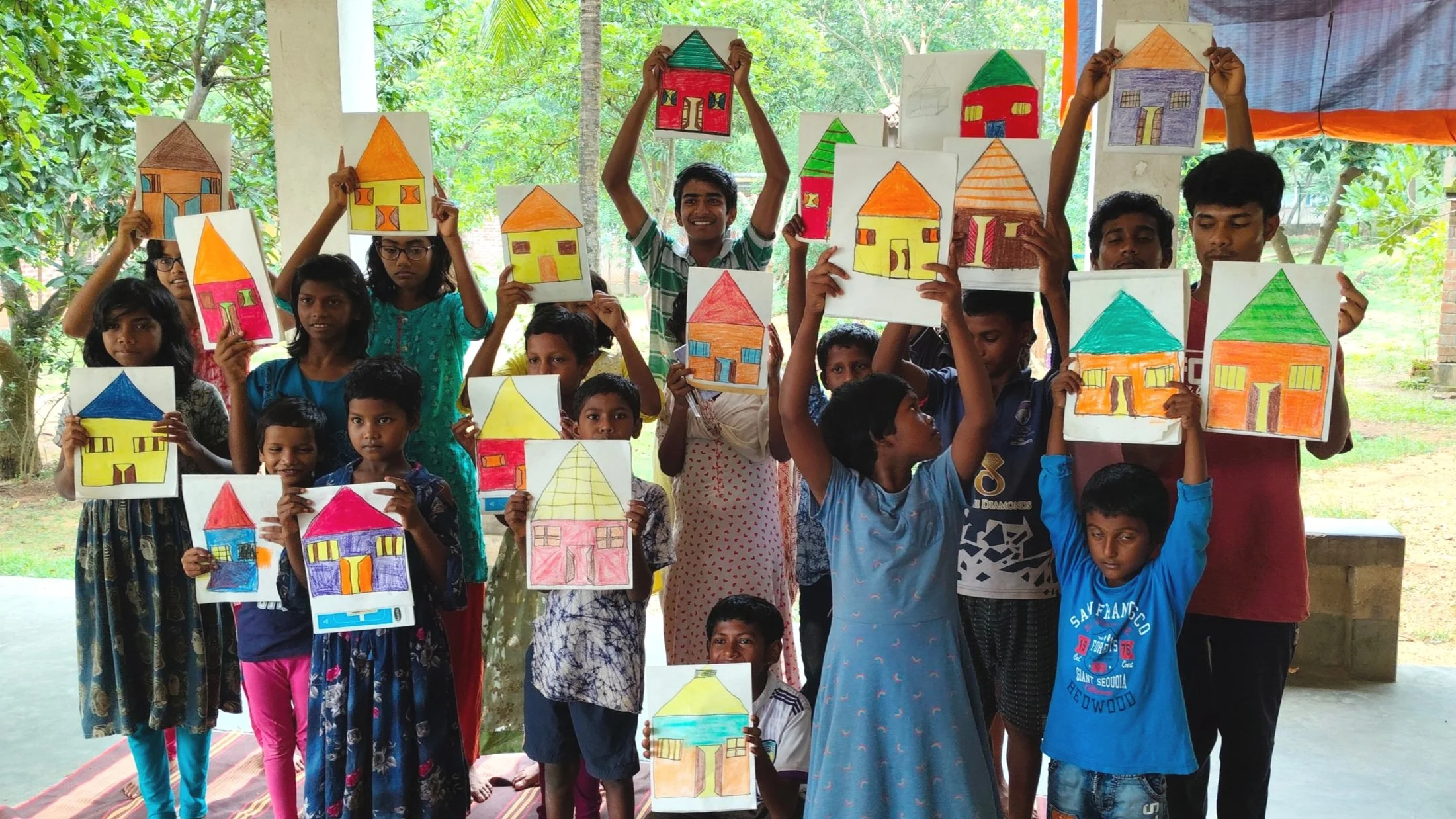 A group of children and a young man holding up colorful drawings of houses, in an outdoor setting with greenery and trees in the background.