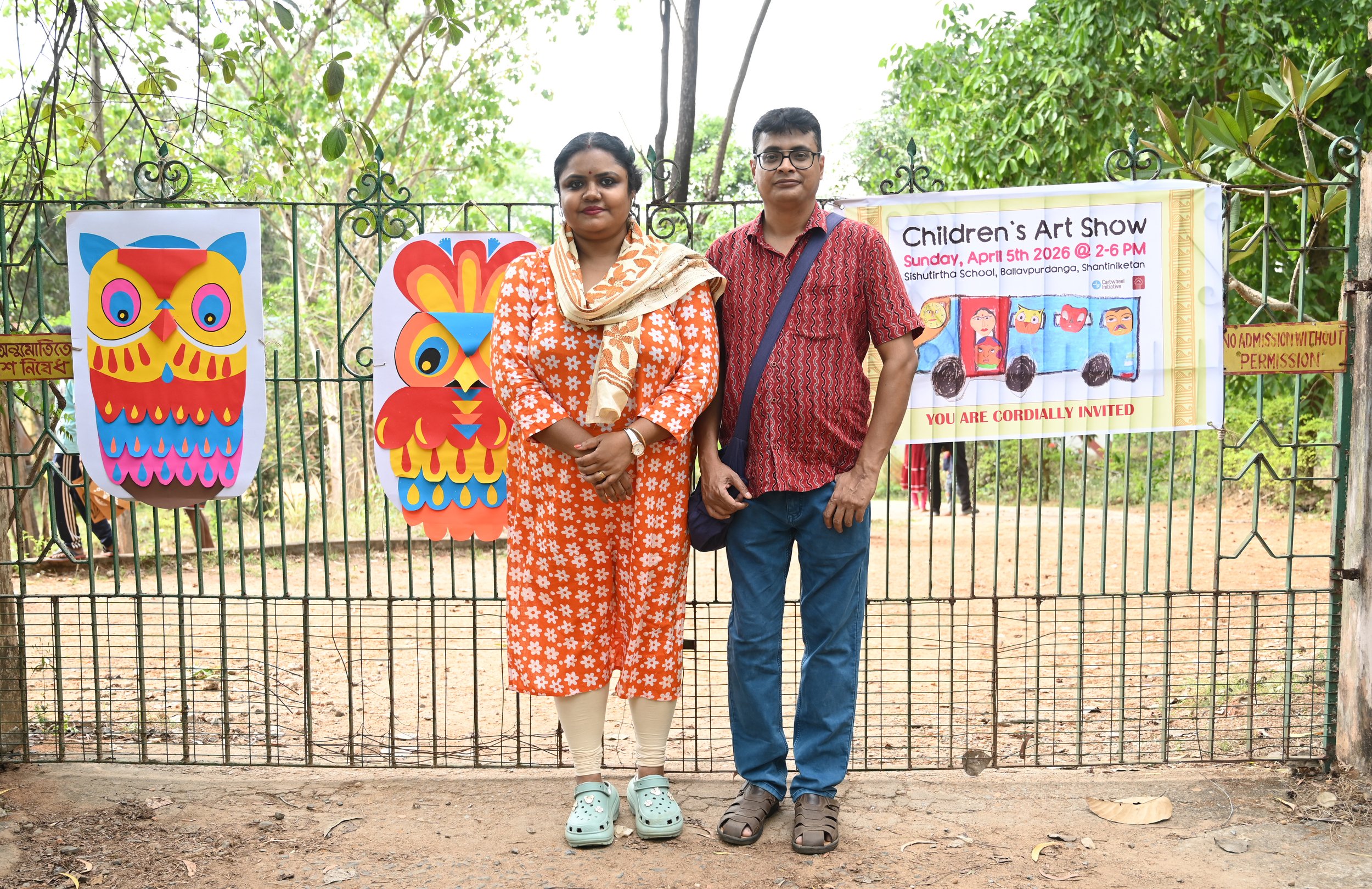 Two adults standing in front of a fence decorated with colorful owl artwork and a banner for a children's art show at Sishurirtha School on April 5, 2026. The woman is wearing a bright orange dress with white floral patterns, cream leggings, and ligh