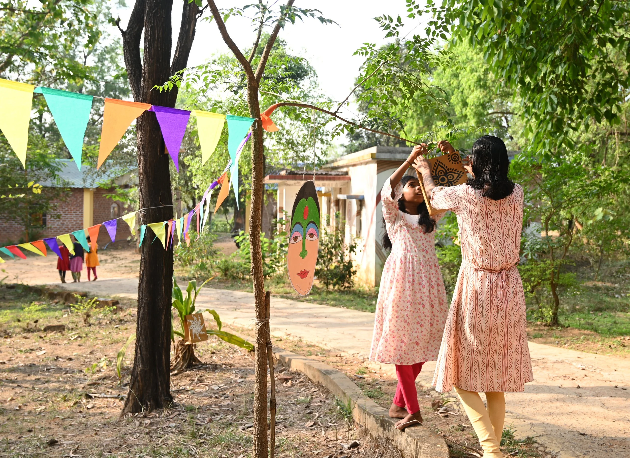 Two women in traditional South Asian attire hanging colorful paper decorations on a tree in a garden area with children playing in the background.