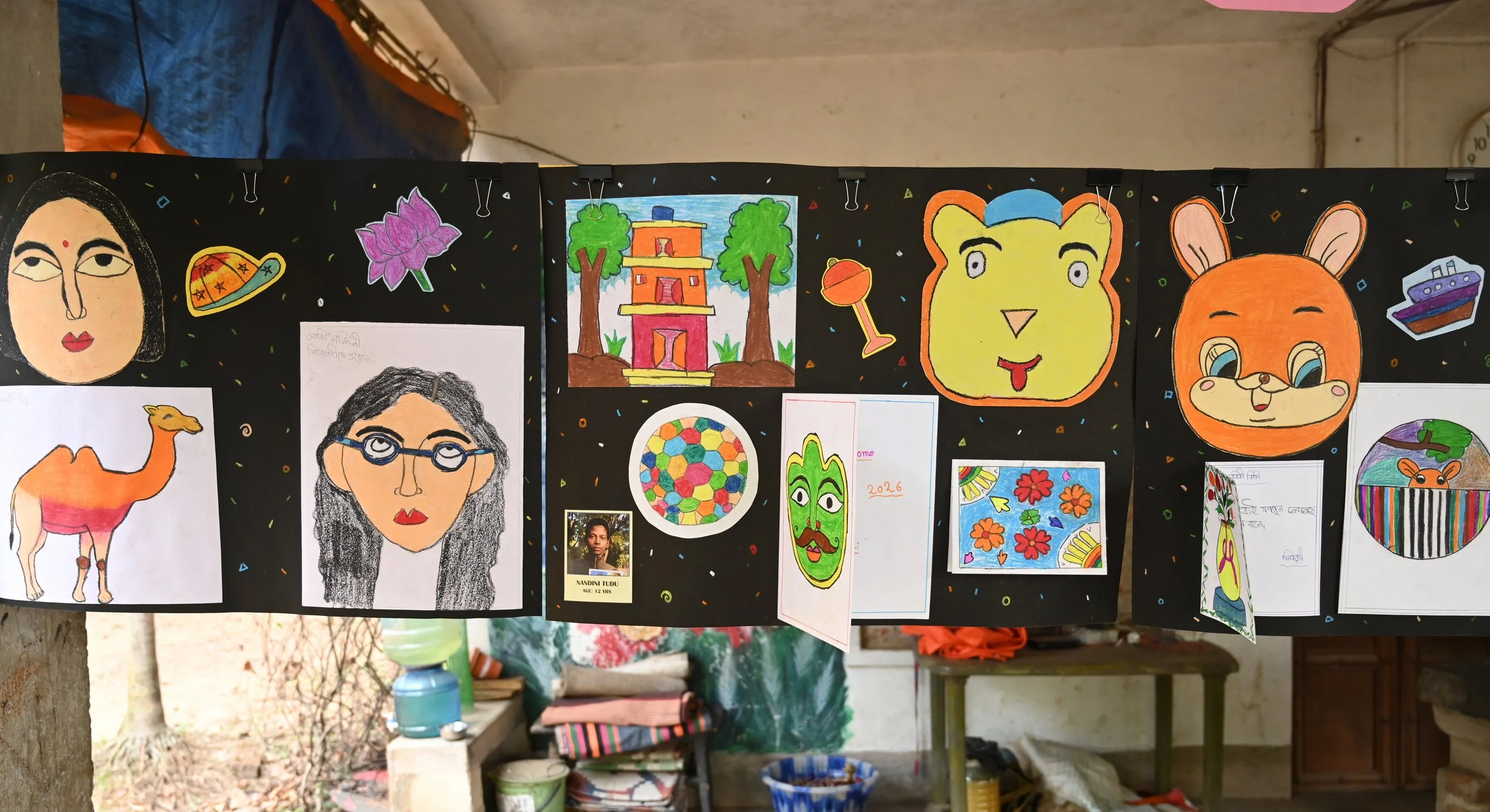 Display of children's colorful drawings and paintings hanging on a black background.