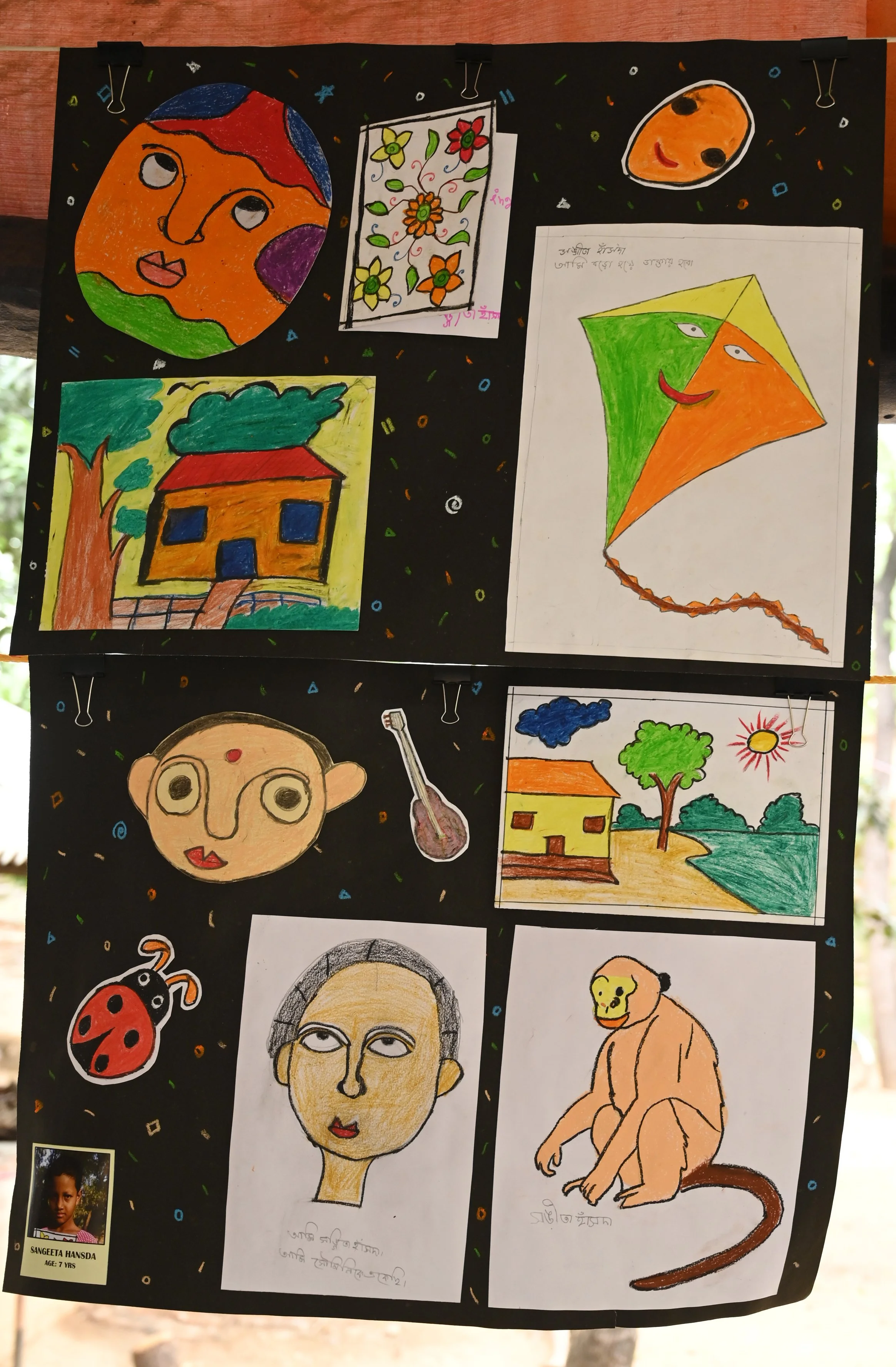 A black display board decorated with colorful children's drawings and paintings, including a face with a map of the world on it, flowers, a kite with a face, a house, a cartoon portrait, a ladybug, a lion, and a boy's photo with the name SANGEETA HAN