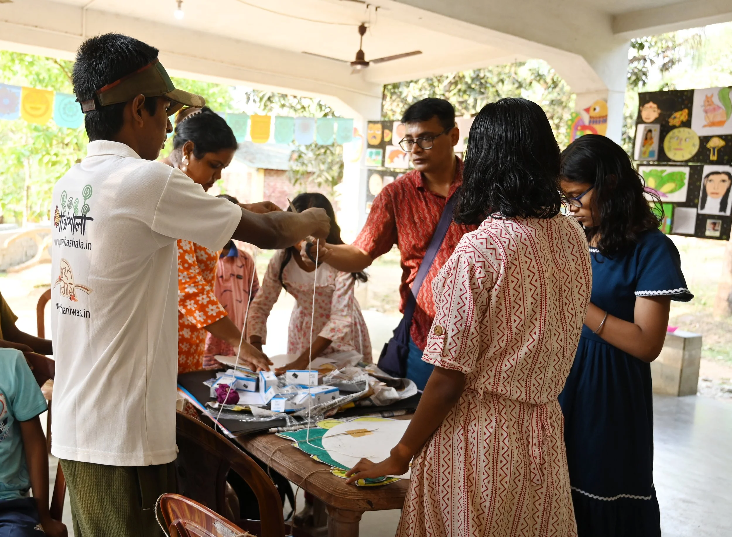 A group of people gathered around a table at an outdoor art or craft exhibition, looking at various items. Some are engaging with the items, and wall art is displayed in the background.