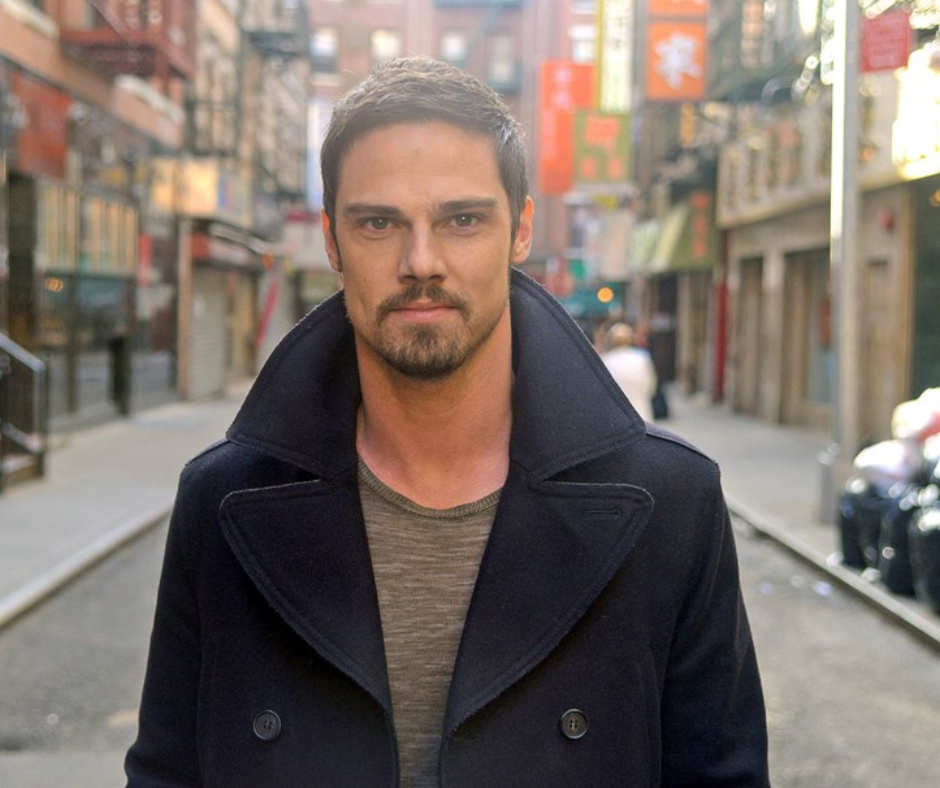 Editorial portrait from a 2018 Who magazine interview: Jay Ryan in a dark peacoat standing in a city street with blurred shop signs.
