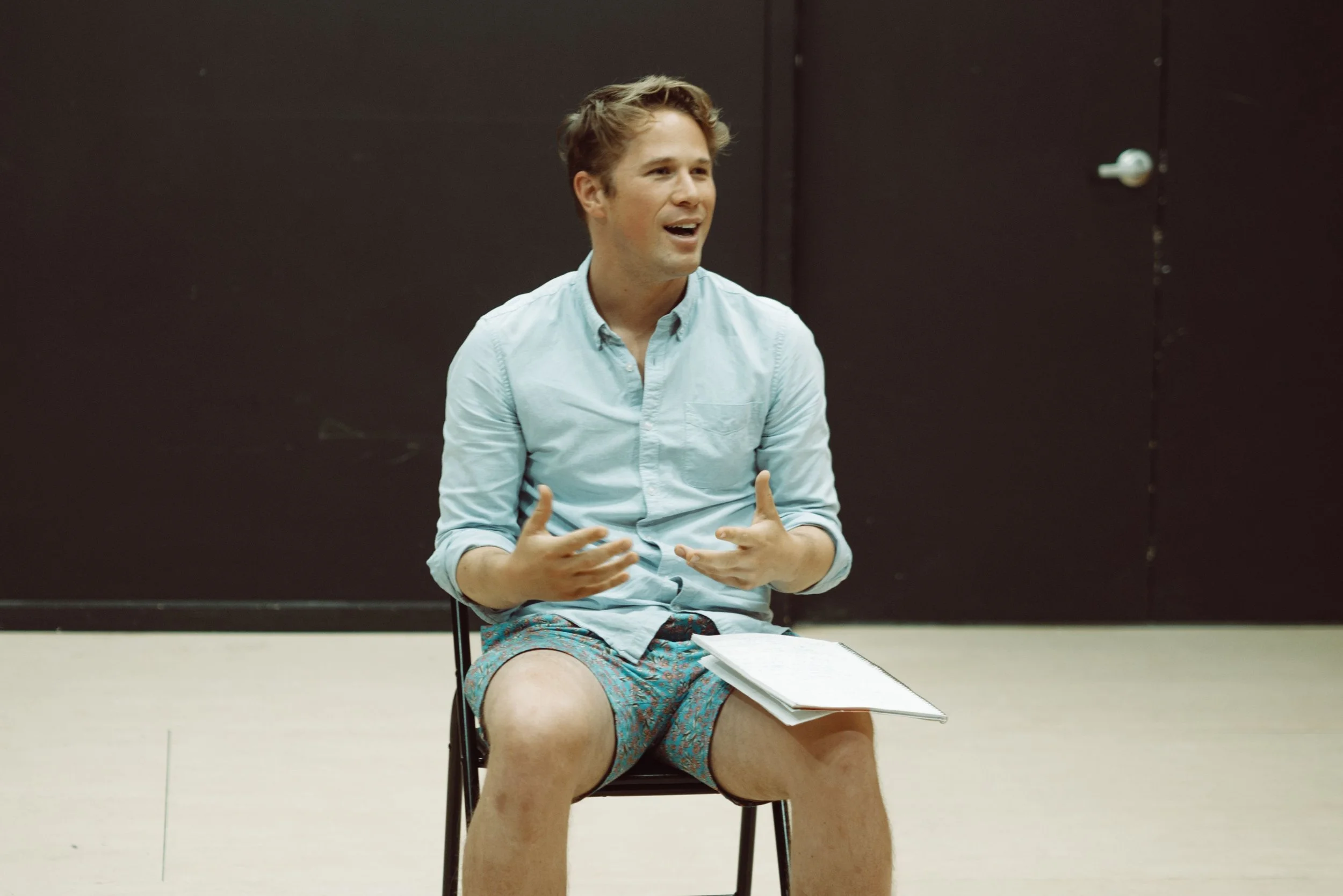 Actor seated in a rehearsal space during a creative development session at Howard Fine Acting Studio, holding a notebook and speaking animatedly while presenting original work.